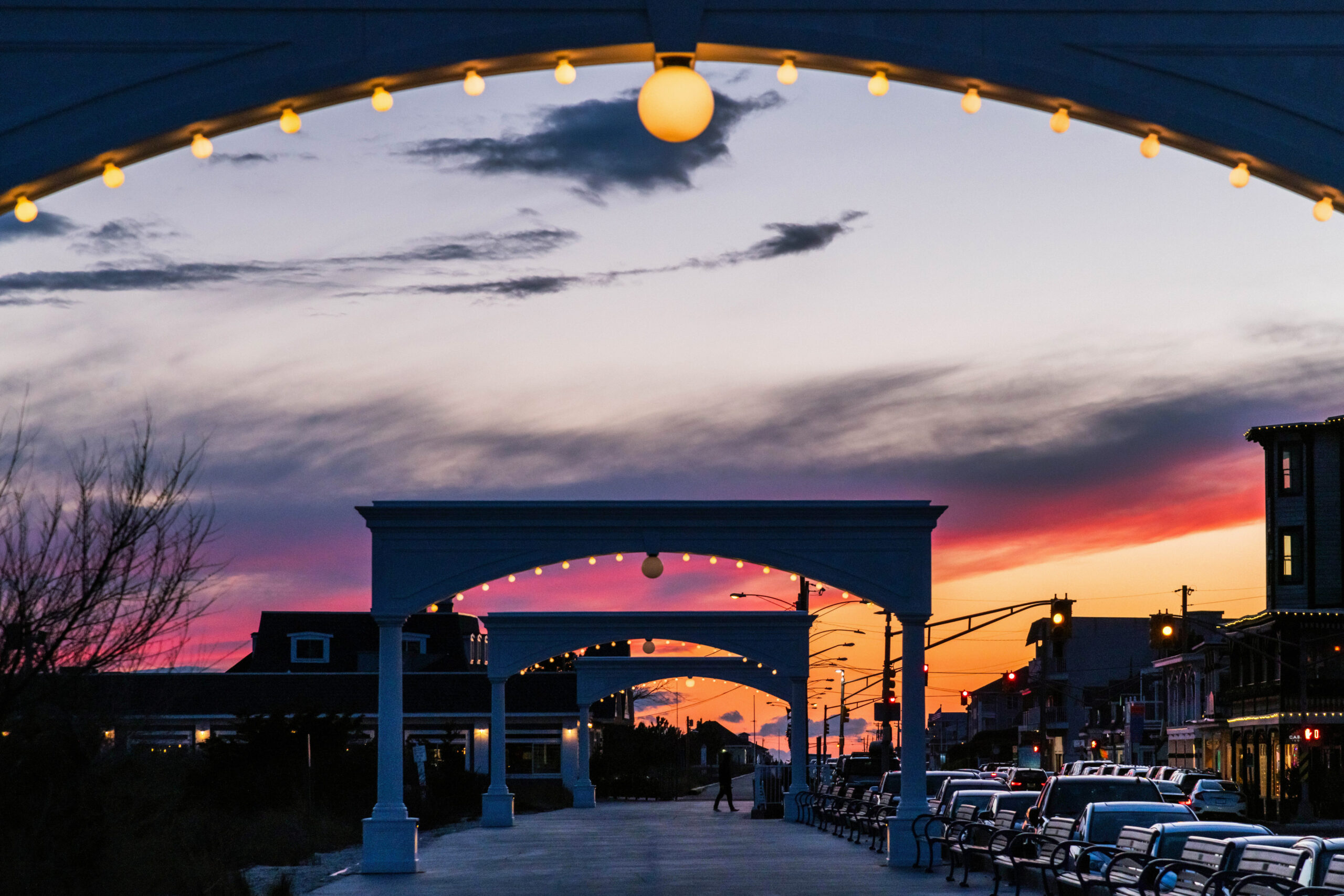 A wide view of the arches on the promenade at sunset. There are colorful purple and pink clouds in the orange and blue sky. The arches are in a row in the center with building on Beach Ave on the right. An arch with lights on is framing the top of the image.