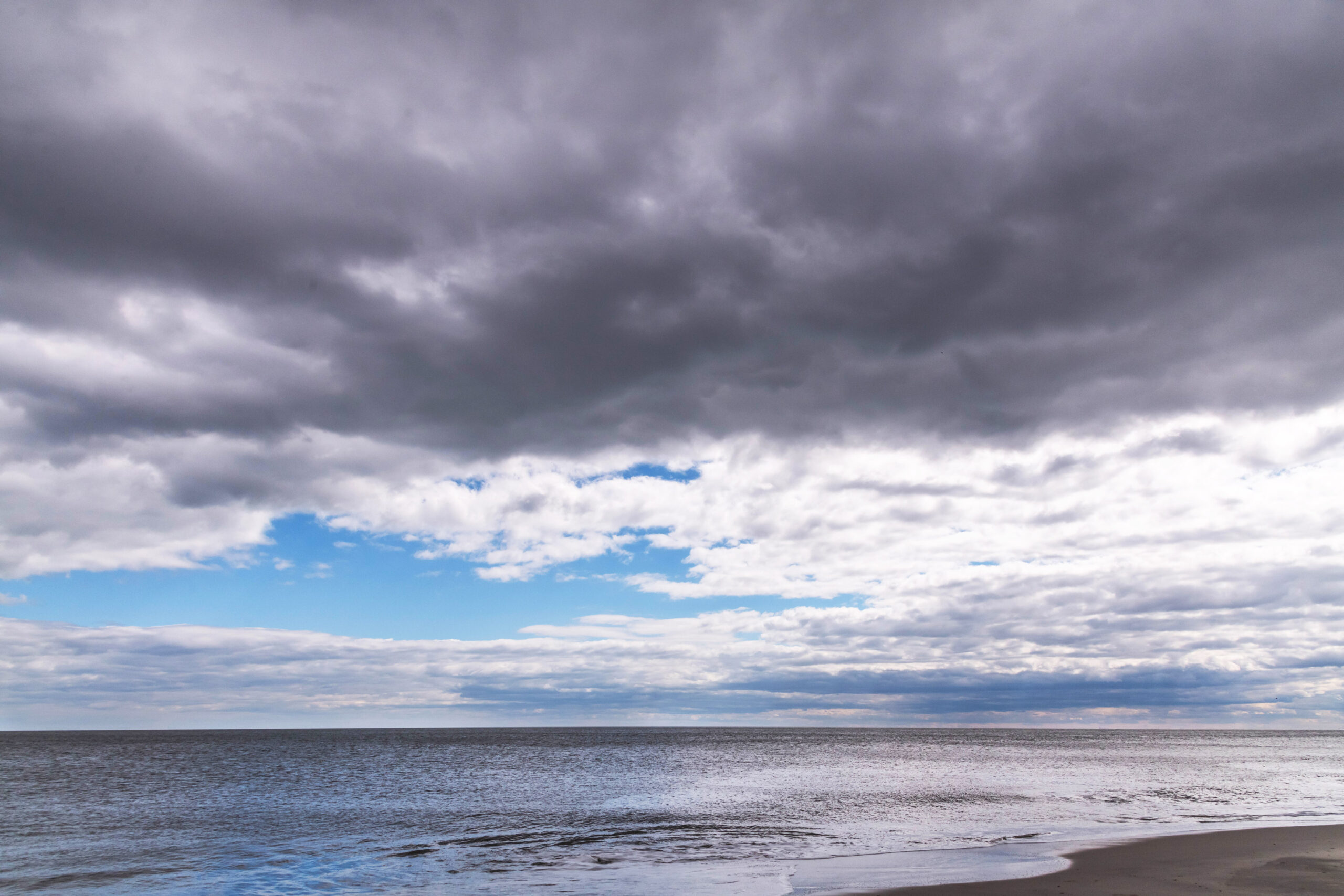 A wide view of the ocean and the sky. There is a big dark cloud with a break of blue sky. More dark thin clouds are at the horizon with white thin clouds. The colors of the clouds are reflected in the calm ocean.