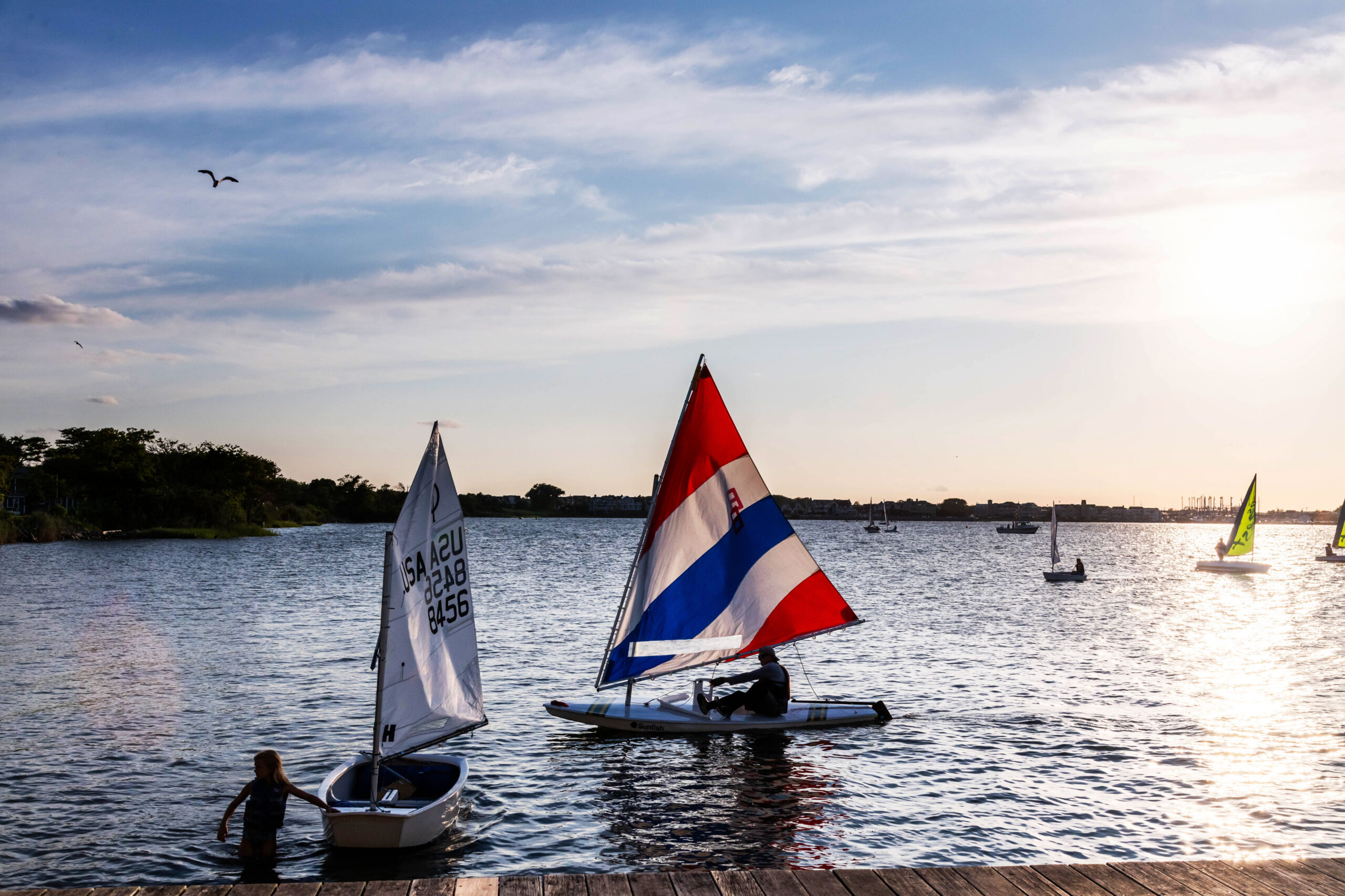 A wide view of racing sailboats in the water. One boat has a red, white, blue, white, and red striped sail. The other sailboat has a white sail with numbers. The sky is blue with thin white clouds, and sunlight is reflected off the water.