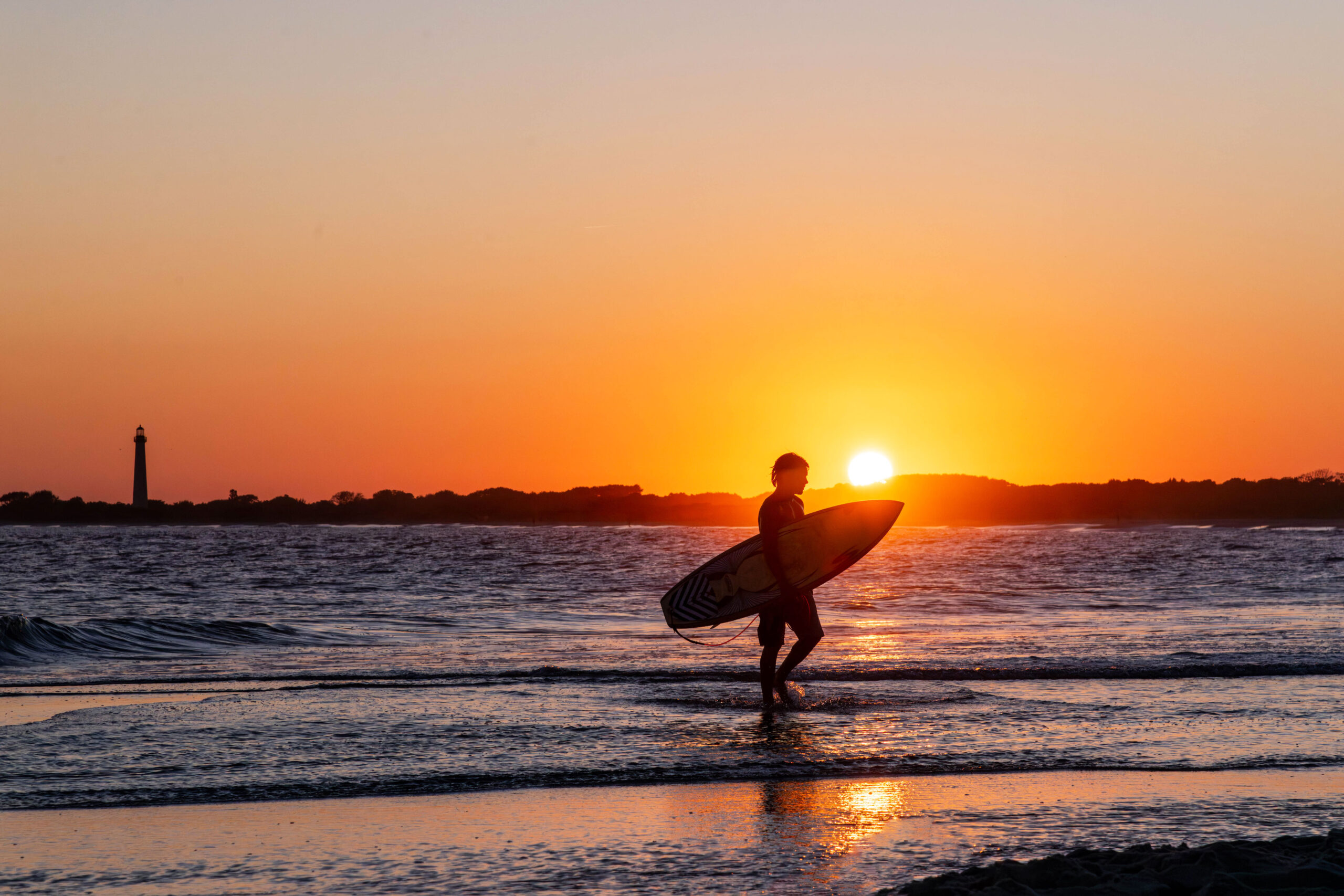 A surfer silhouetted by the setting sun, walking out of the ocean. The Cape May Lighthouse is in the distance, and the sky is clear and golden.