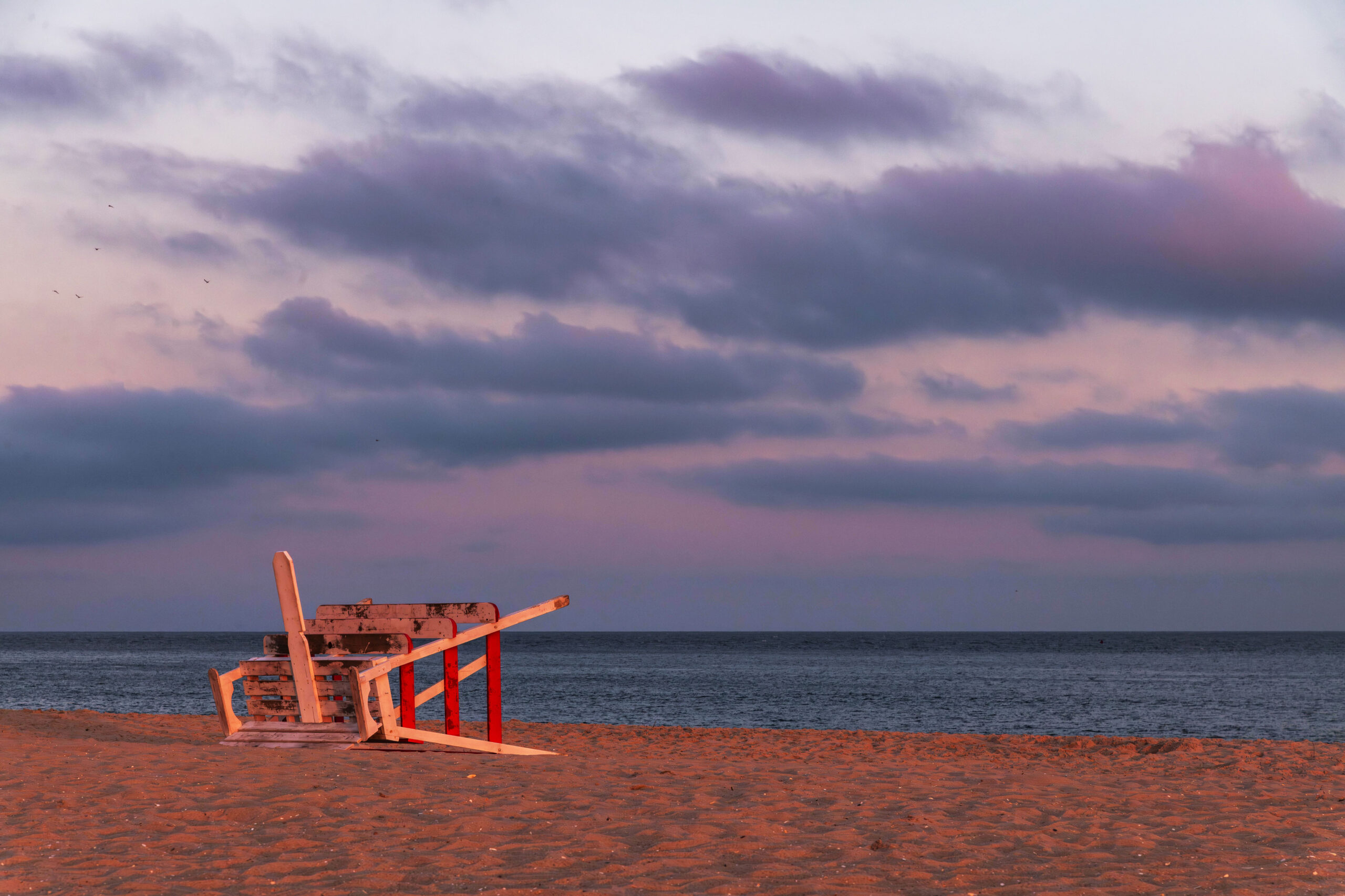 A red and white Cape May lifeguard stand down on its side on the beach at sunset. There are purple and pink clouds in the sky, and golden light is shining on the stand and the sun.