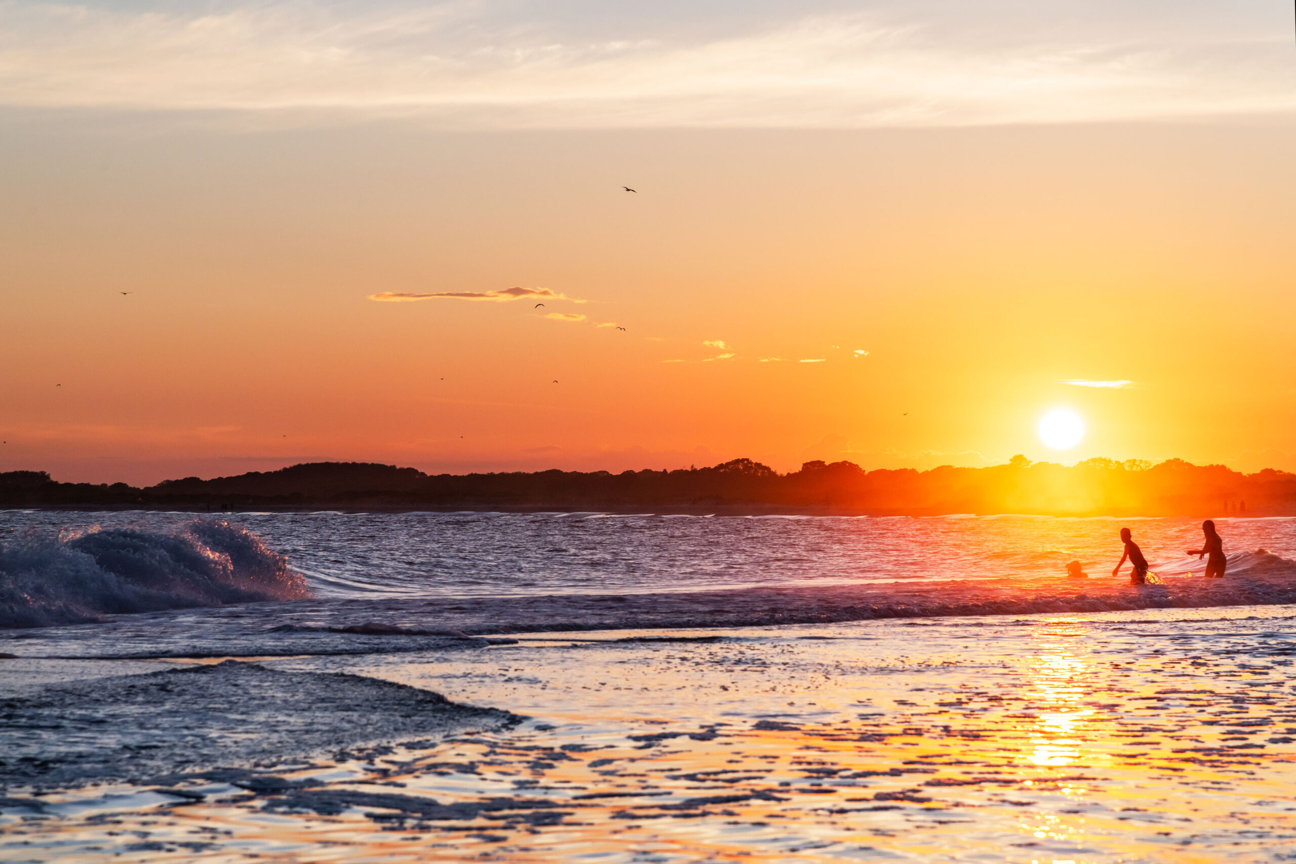 A wide view of sunset at the ocean. The water is close in the foreground with a wave crashing. The setting sunlight is shining on the water. Three people are silhouetted in the ocean.