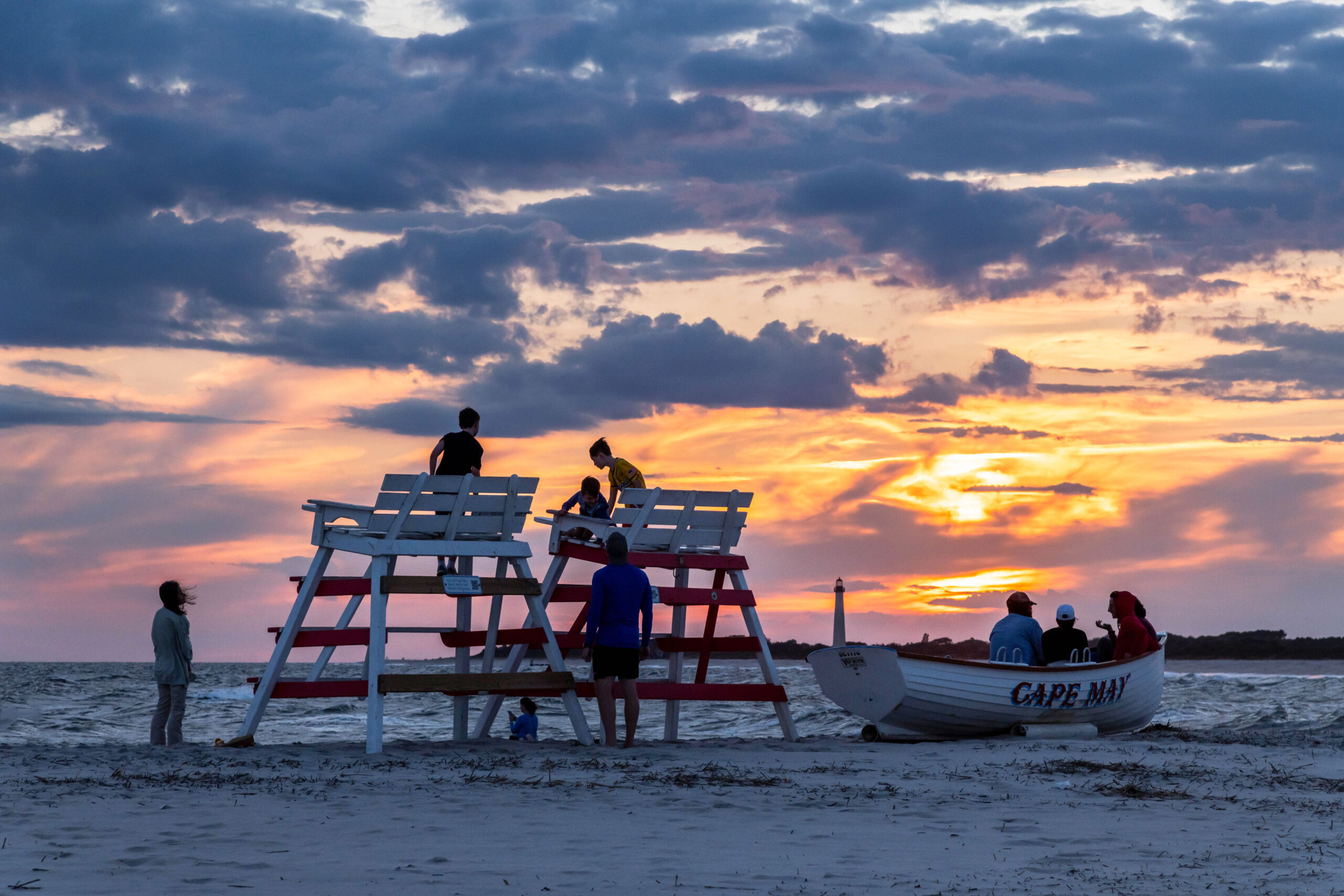 A wide view of sunset at the Cove beach. A group of people are sitting in a Cape May lifeguard boat. There are two lifeguard stands next to each other, and a family is sitting on them and standing around them. There are puffy purple clouds in the sky, and thinner purple clouds at the horizon. The sky is orange and pink behind the clouds. The Cape May Lighthouse is in the distance.