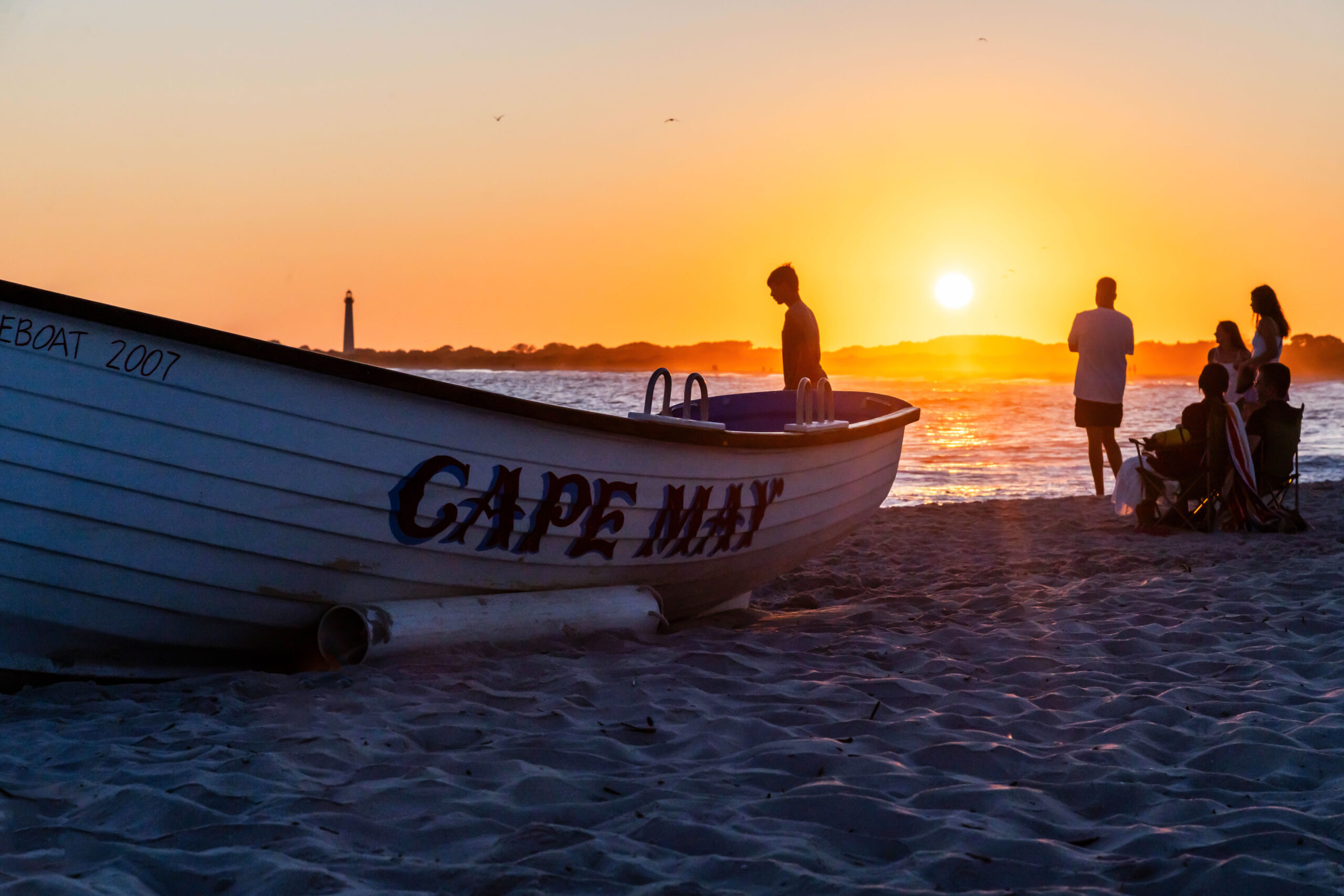 A wide view of sunset at the beach with a Cape May lifeguard boat in the foreground and the Cape May lighthouse in the background. There are a few people silhouetted by the ocean watching the sunset. The sky is clear and golden.