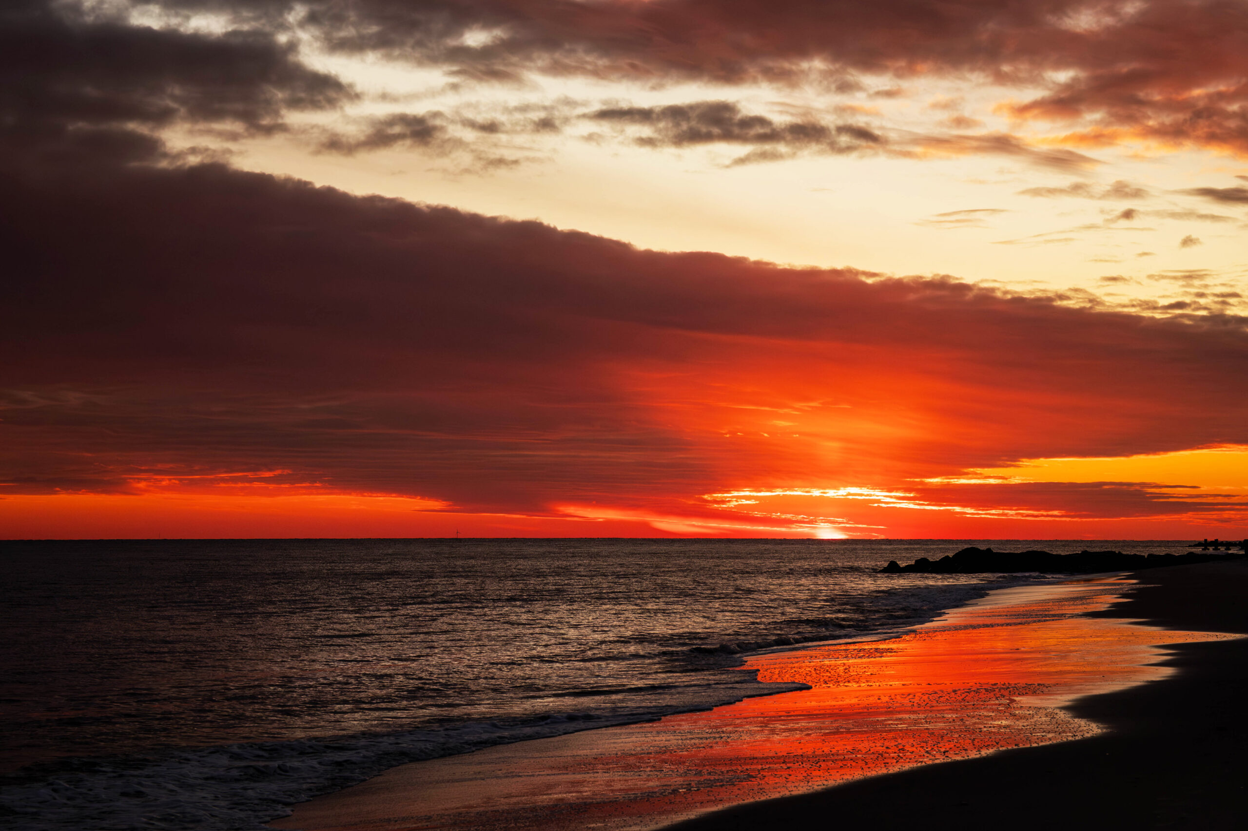 A wide view of a colorful bright sunset over the ocean. There are thin dark clouds in the sky, with bright orange light shining from the sun just setting below the horizon. The orange light is reflected in the shoreline.