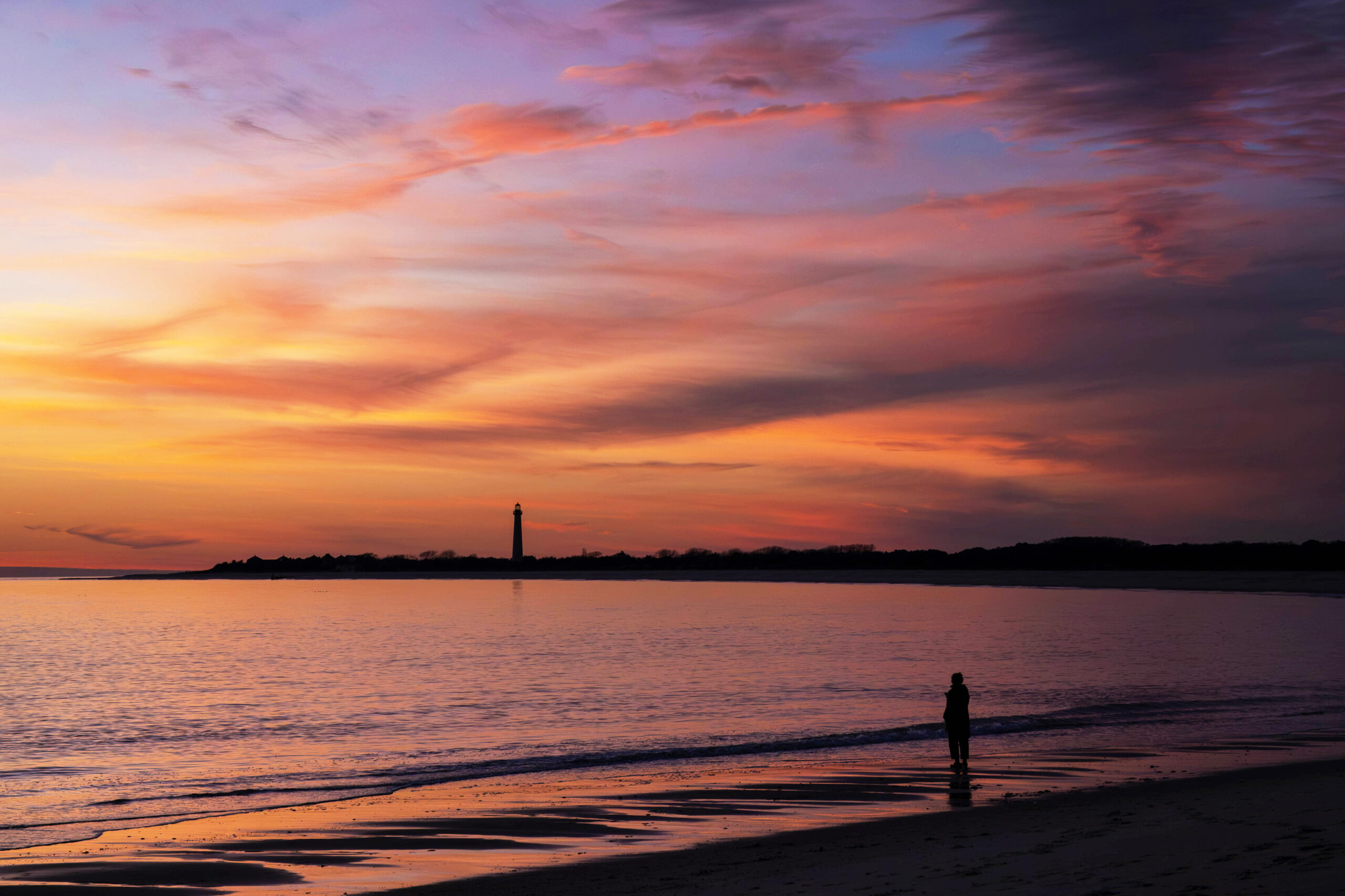 A wide view of a colorful sunset at the beach and ocean. There are thin pink, purple, and yellow clouds in the sky. The colors are reflected in the ocean. Someone is silhouetted standing by the edge of the water. The Cape May Lighthouse is in the distance at the horizon line.