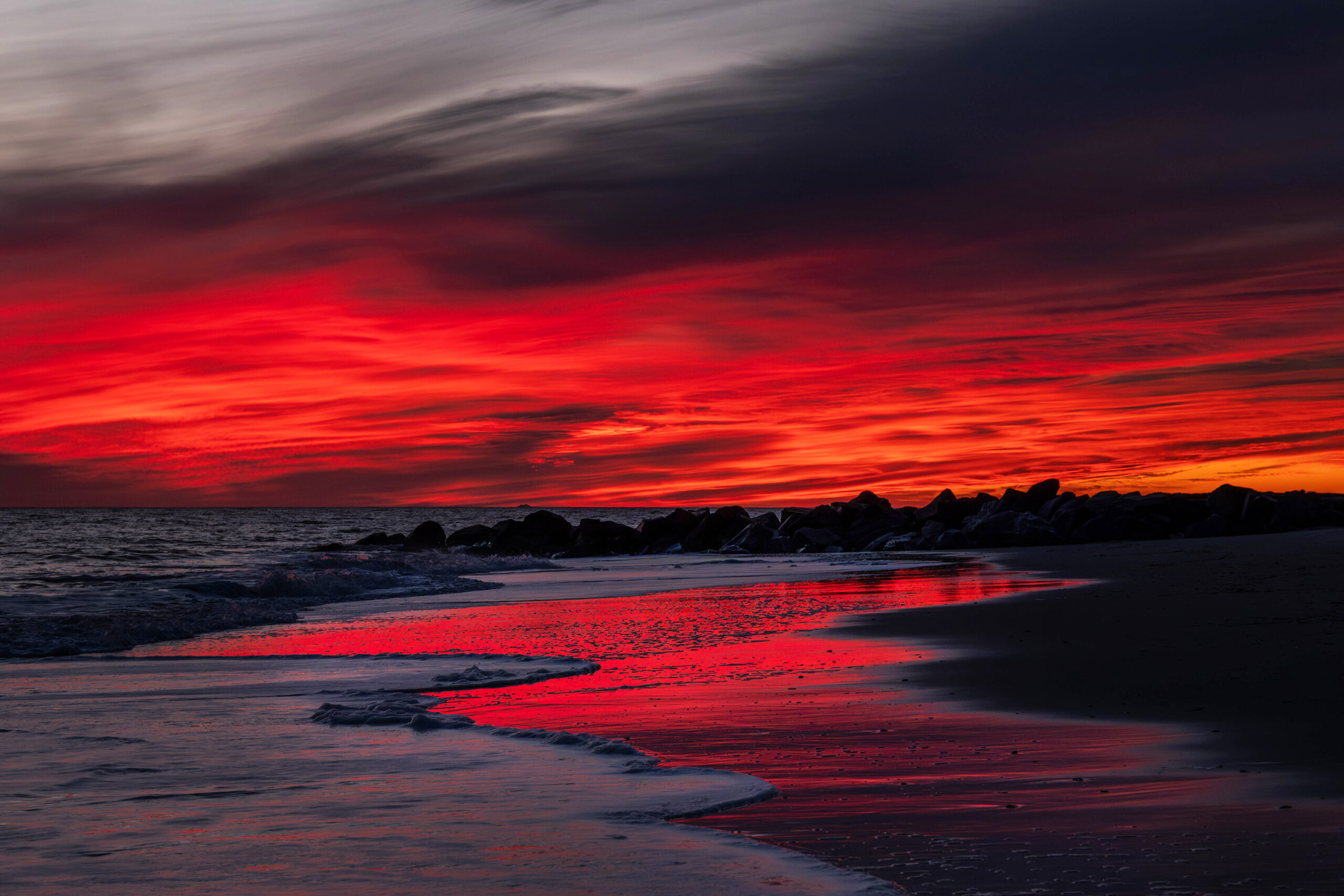 A wide view of a colorful sunset at the beach. There are red and dark purple bright clouds at the horizon. The red is reflected in the shoreline next to the blue ocean.