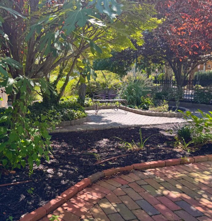 view through light dappling to a wrought iron butterfly bench on brick pathway