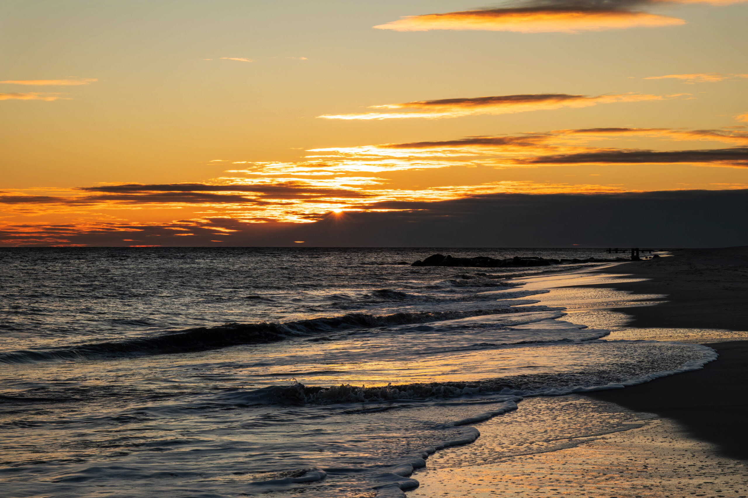 A wide view of the sunset over the ocean. The sun is setting behind thin purple clouds at the horizon. The sky is orange with a few small lines of clouds in the sky. The ocean is yellow and blue, and the sunlight is reflected in the shoreline.