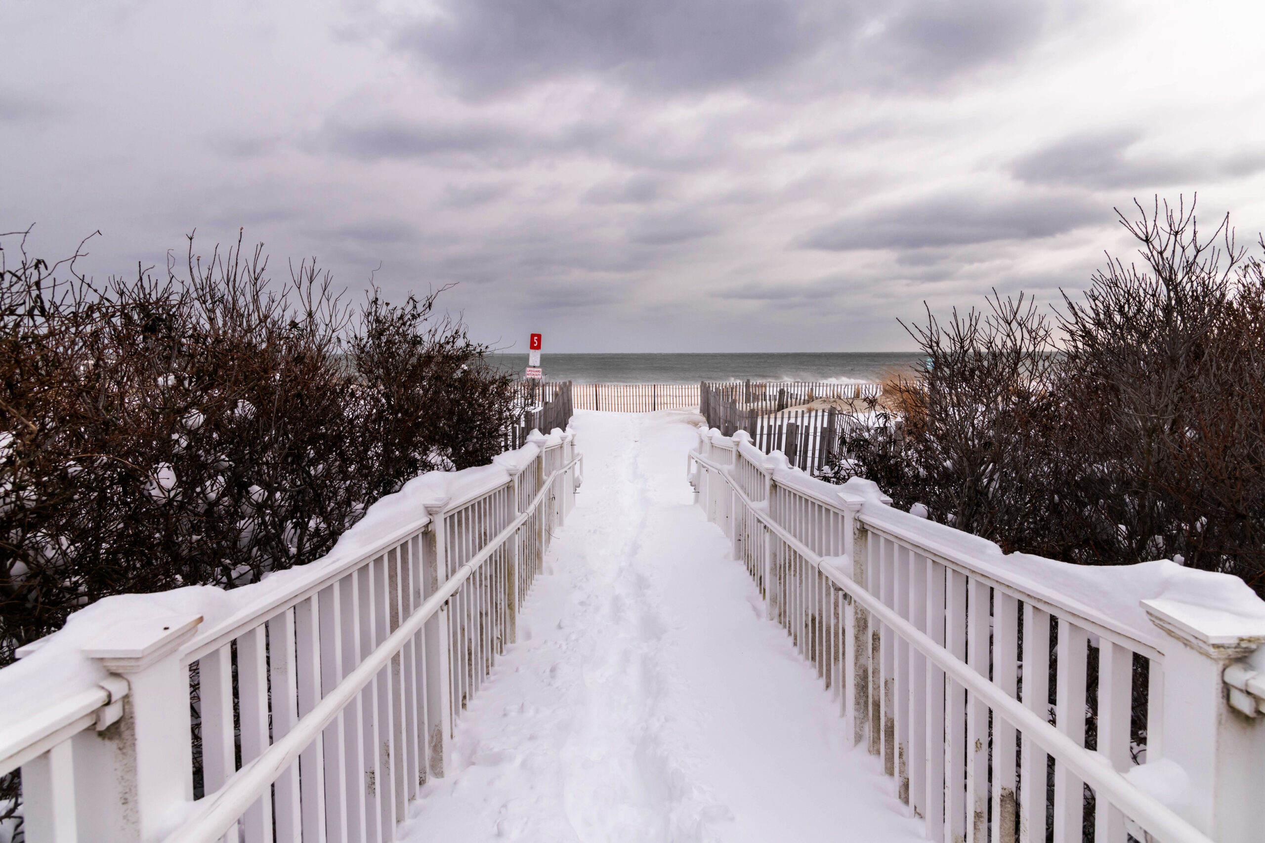 A wide view of a fenced path to the beach covered with snow and gray clouds in the sky.