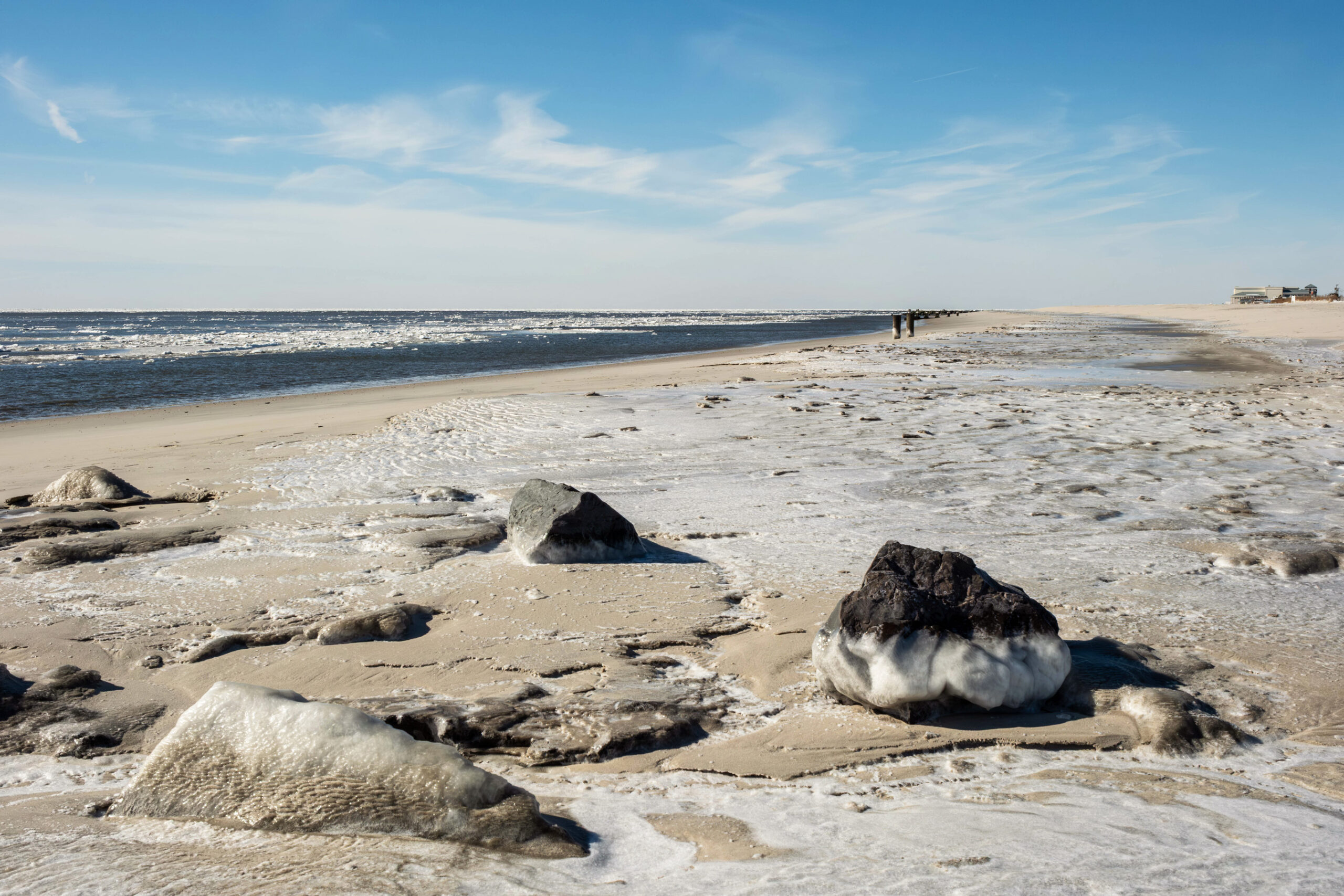 A wide view of ice and frozen snow on the beach with blocks of ice in the ocean. There are a few rocks in the foreground with ice on them. The sky is blue with a few thin clouds.