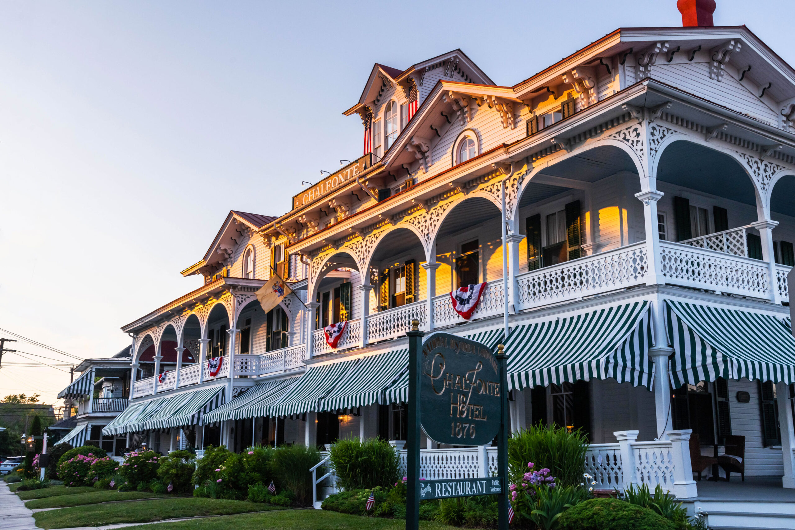 A wide view of the Chalfonte at sunset. Sunlight is shining on the top of the building, while the bottom half of the building is in shadow. There are green and white awnings on the first floor porch, and a few American flag buntings are hanging on the second floor porch.