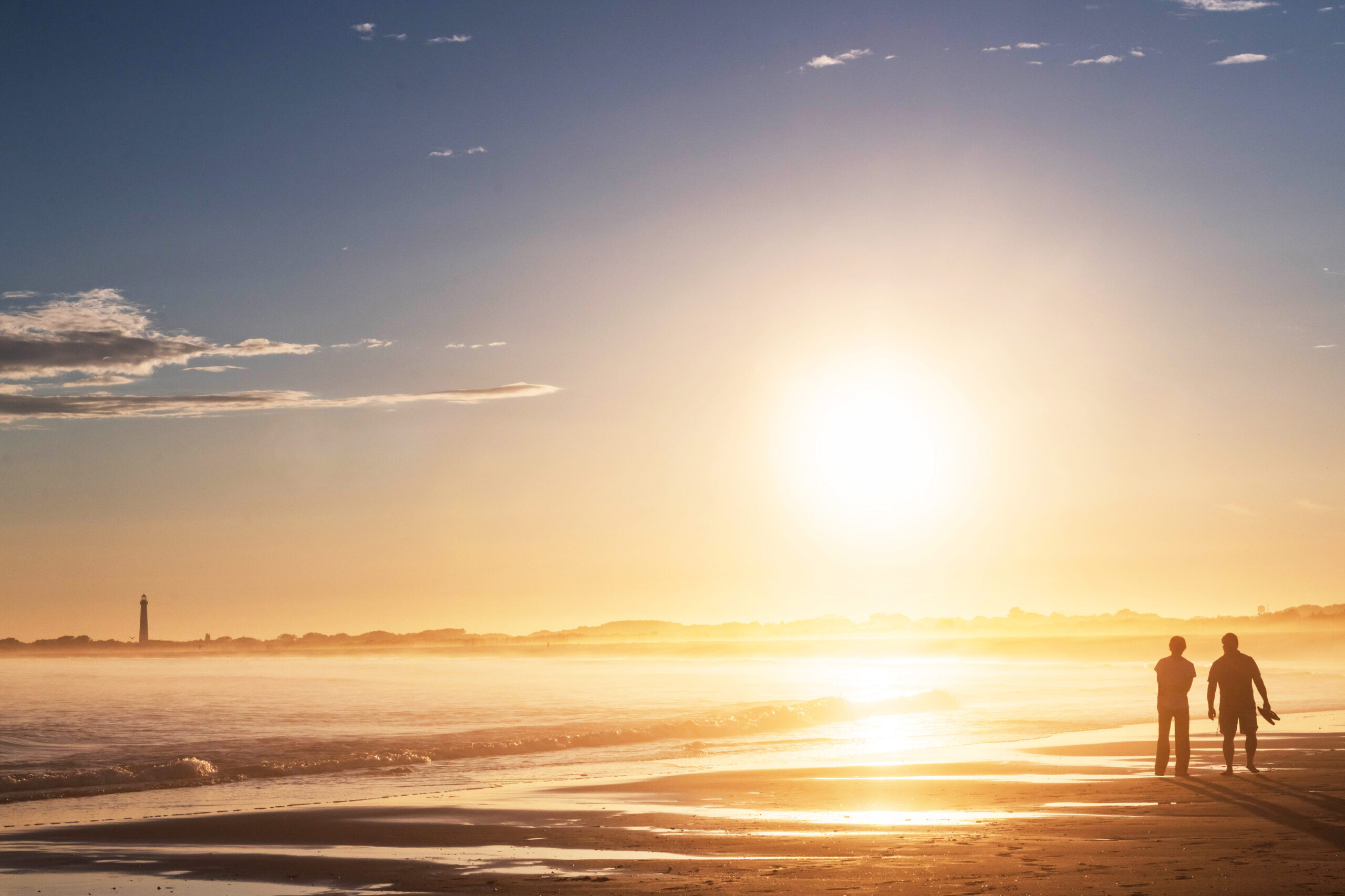 Two people walking on the beach with the Cape May Lighthouse in the distance and the sun shining bright.