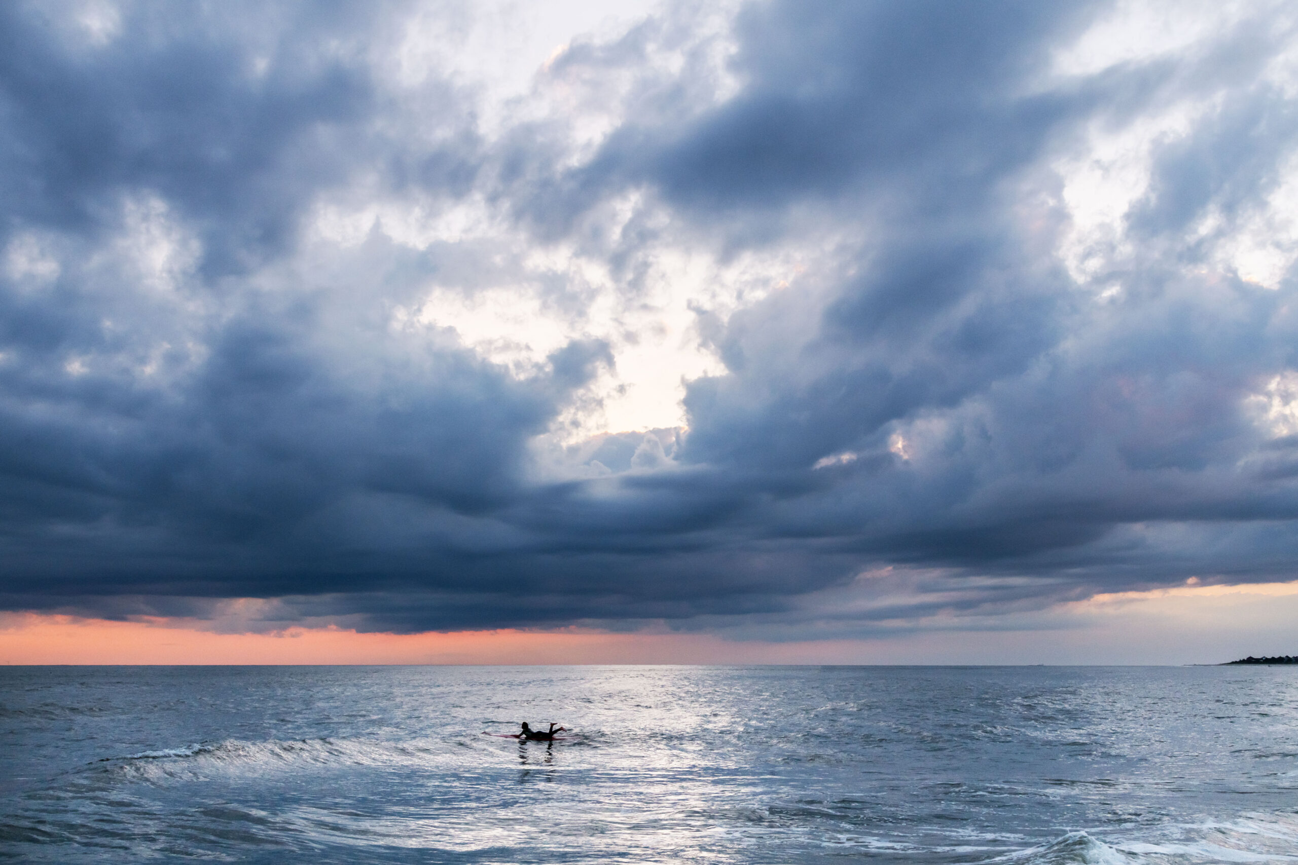 A wide view of the sky and ocean. big dark blue and purple clouds are in the sky. There is pink at the horizon, and some clear sky breaking up the clouds. A lone surfer is paddling in the ocean.