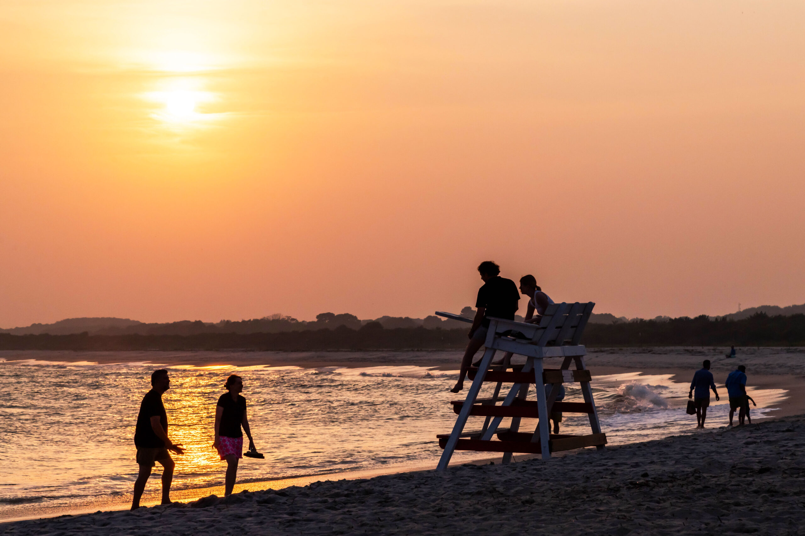 A wide view of people at the beach at sunset. The people are all silhouetted by the setting sun. There is a couple walking in the foreground, another couple sitting on a lifeguard stand, and another group walking in the distance by the ocean. The sky is orange and yellow.