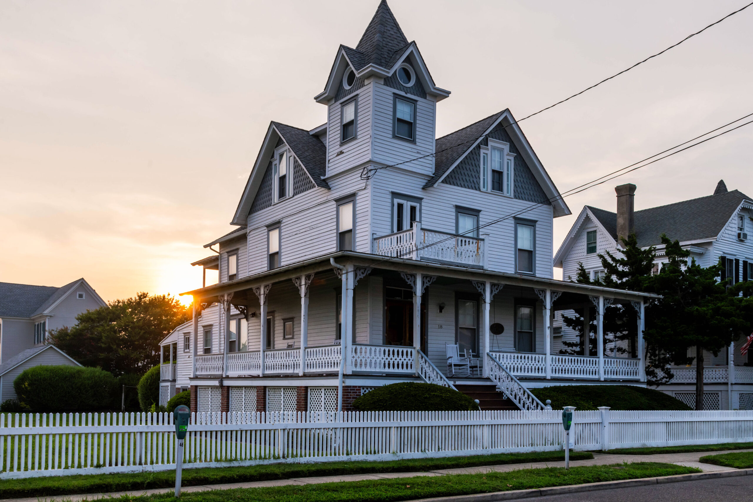 A wide view of a Victorian style house. The house has white siding and a white picket fence. The sun is setting behind the house, and the sky is yellow and white.