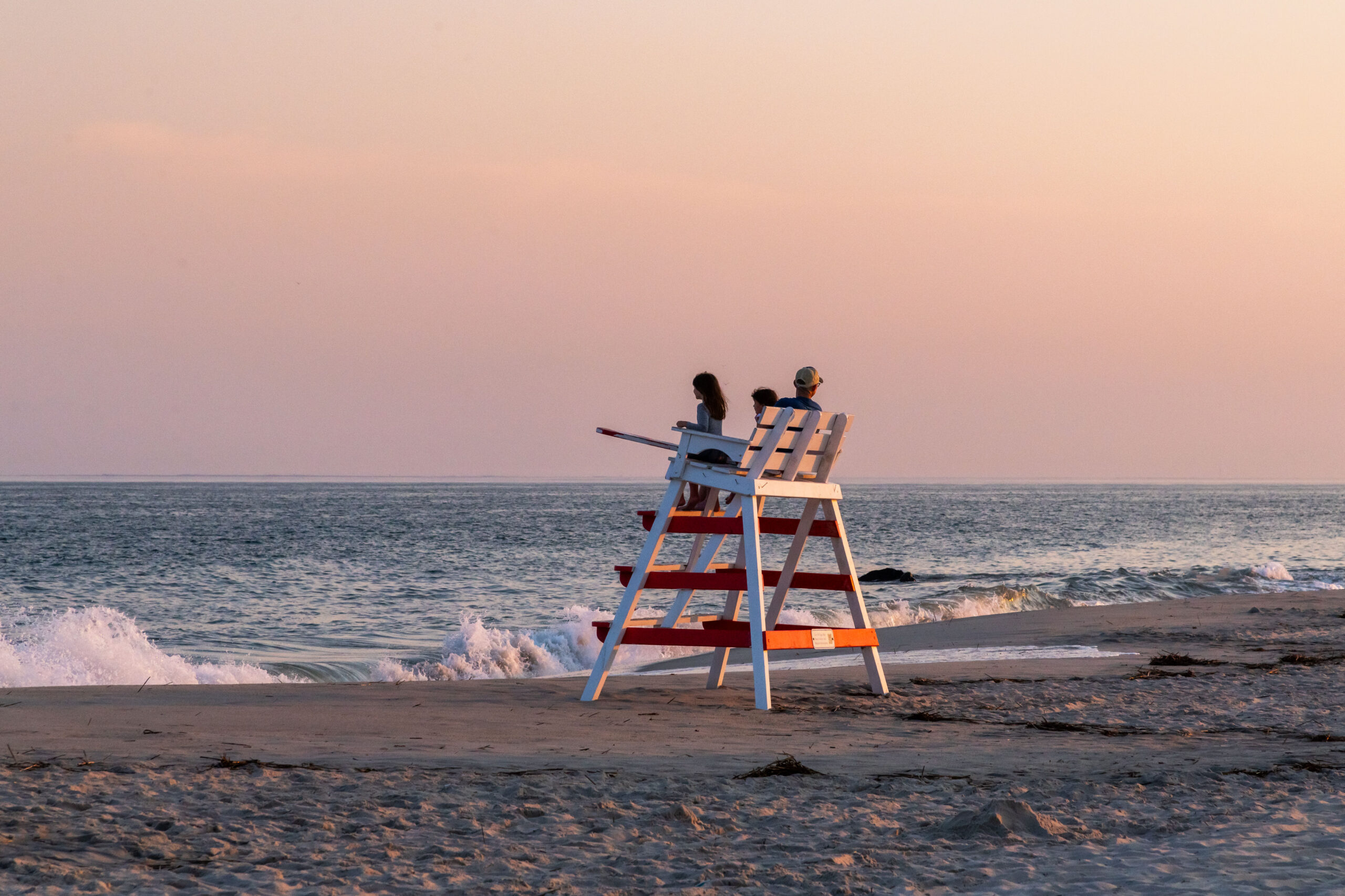 A man and two children sitting on a lifeguard stand at the beach at sunset. A wave is crashing in the ocean. The sky is light pink and orange.