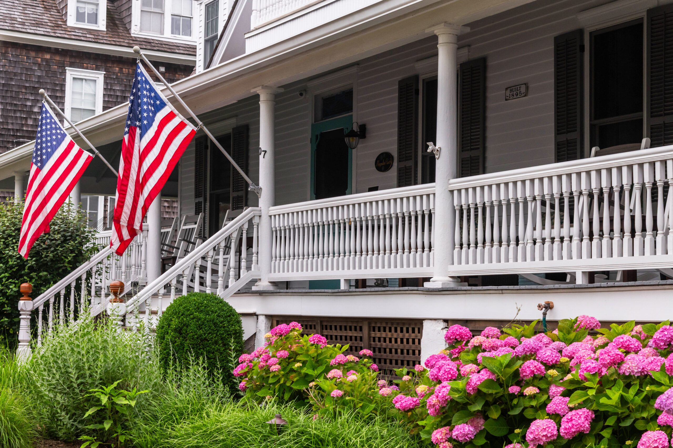 A wide view of a white front porch with two American Flags and pink hydrangeas.