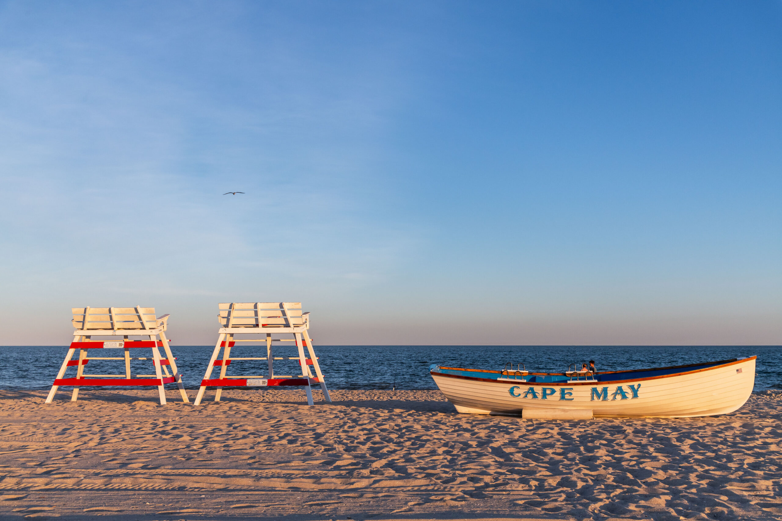 Two lifeguard stands an a Cape May lifeguard boat on the beach. The lifeguard stands are red and white. The boat is white with blue writing. There is a bright blue sky with a few wispy thin clouds on the left. One seagull is flying in the sky, and the sun is shining on the beach.