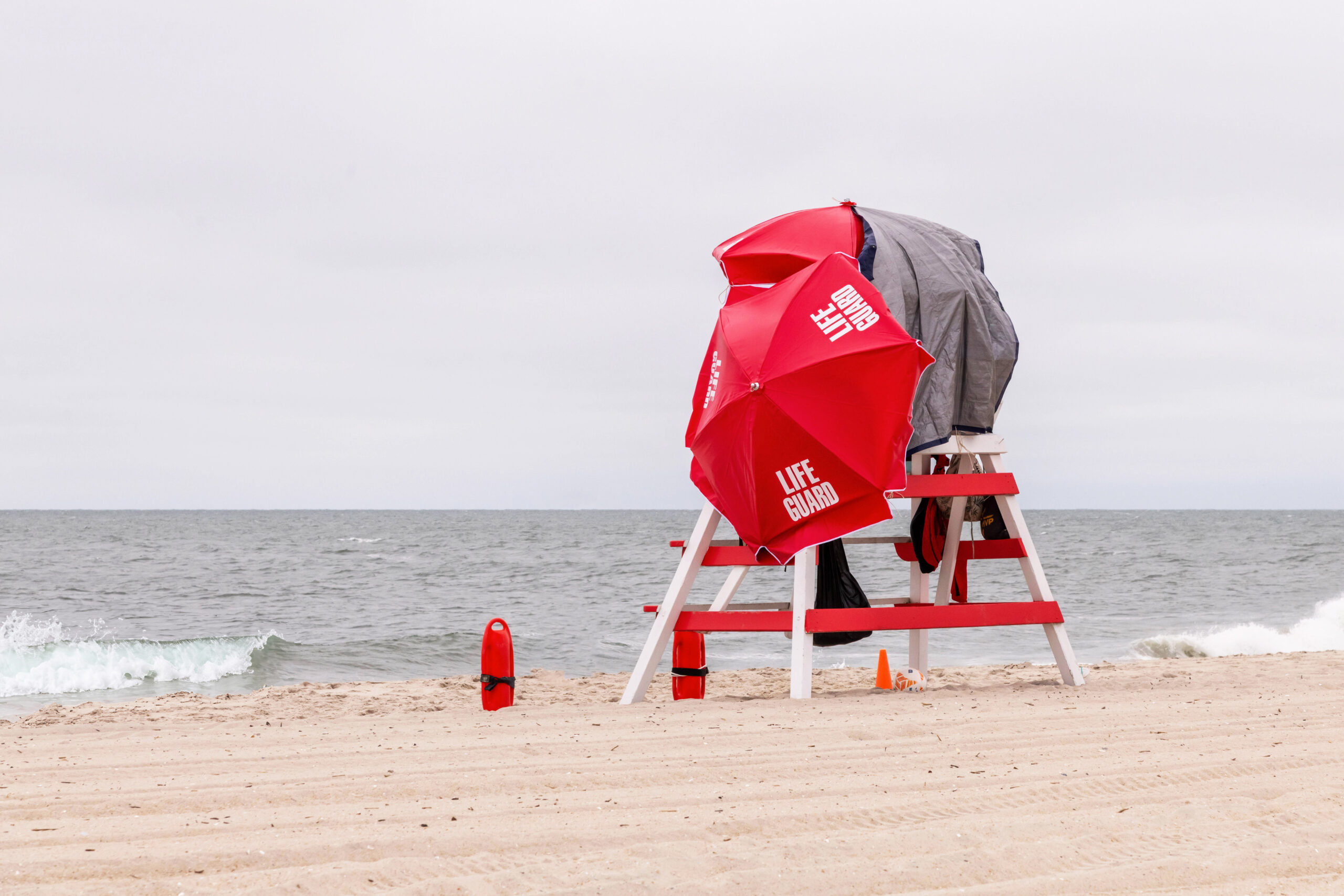 A wide view of a lifeguard stand on the beach with a cloudy sky. There are two red umbrellas create a wind shield at the top of the lifeguard stand. One red umbrella has white text that says "LIFEGAURD". There is a gray towel covering the back of the stand. There are waves crashing in the ocean, and two lifeguard rescue buoys are sticking upright in the sand by the lifeguard chair.