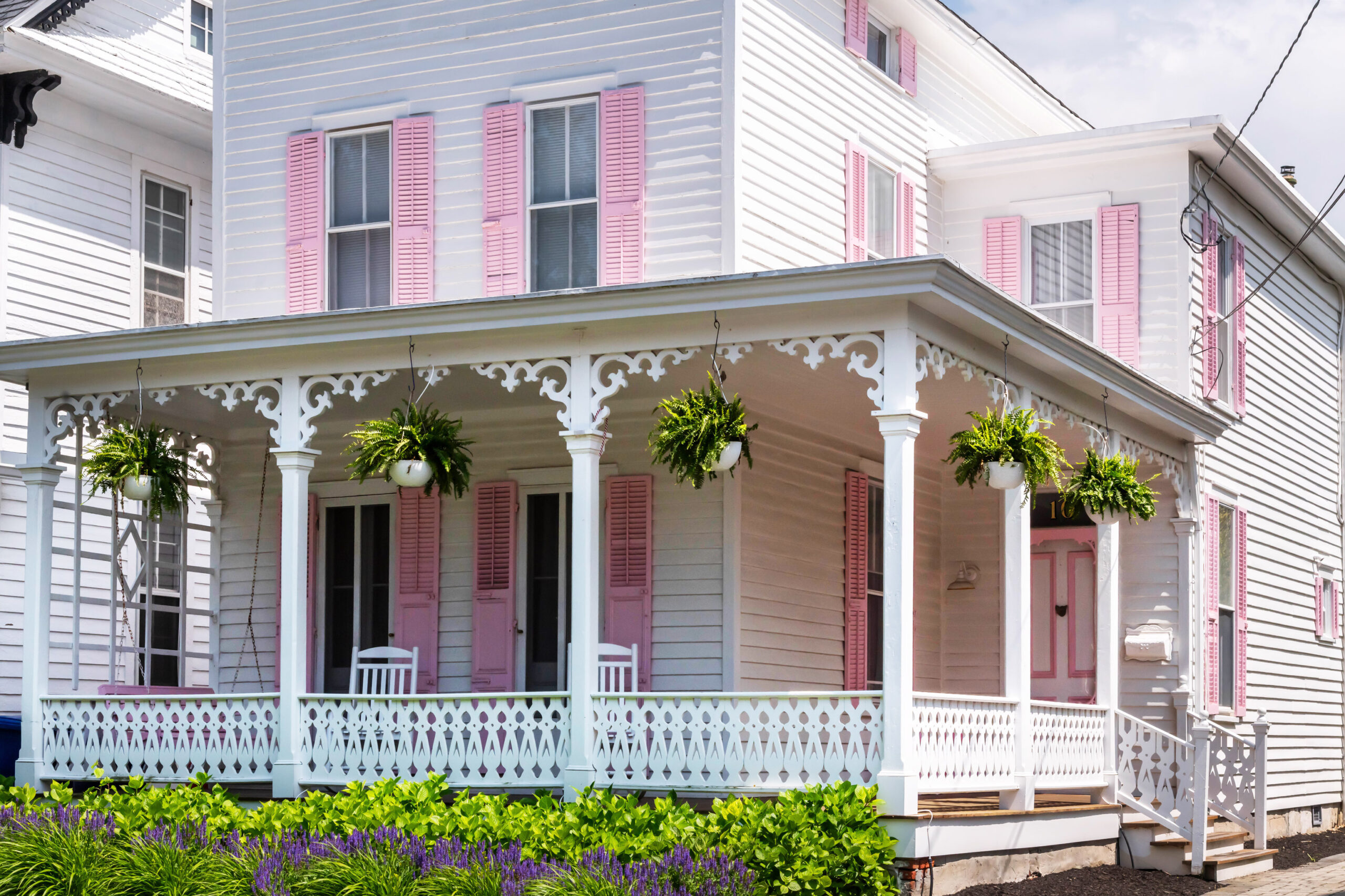 A white and pink Victorian house and front porch. There are green fern plants hanging on the front porch, and green shrubs and purple flowers in front of the porch.