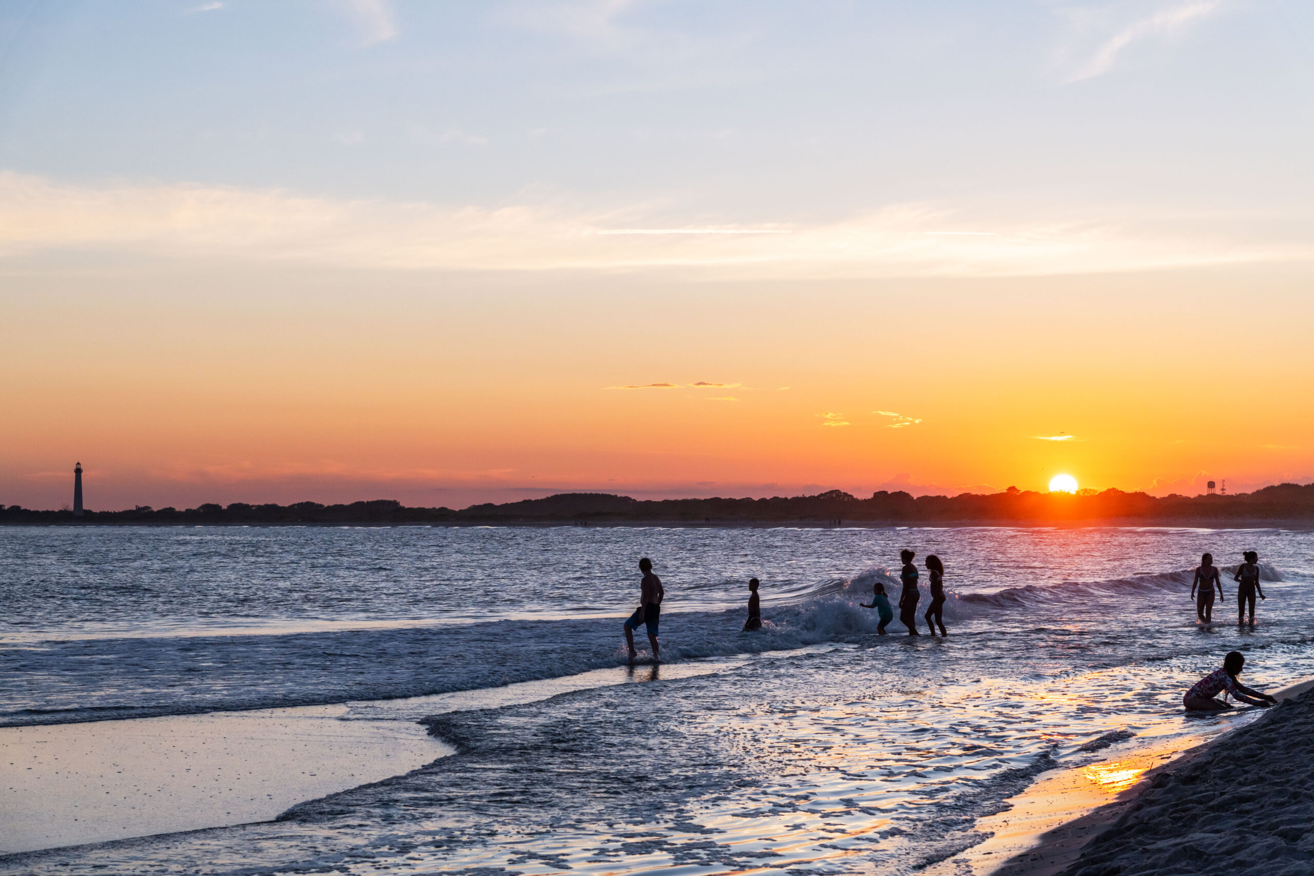 A wide view of sunset at the Cove beach. People are silhouetted walking into the ocean, and playing in the water as waves rush into the shore. The Cape May Lighthouse is in the distance at the horizon. There is a clear blue, orange, and pink sky, and the sun is dipping below the horizon.