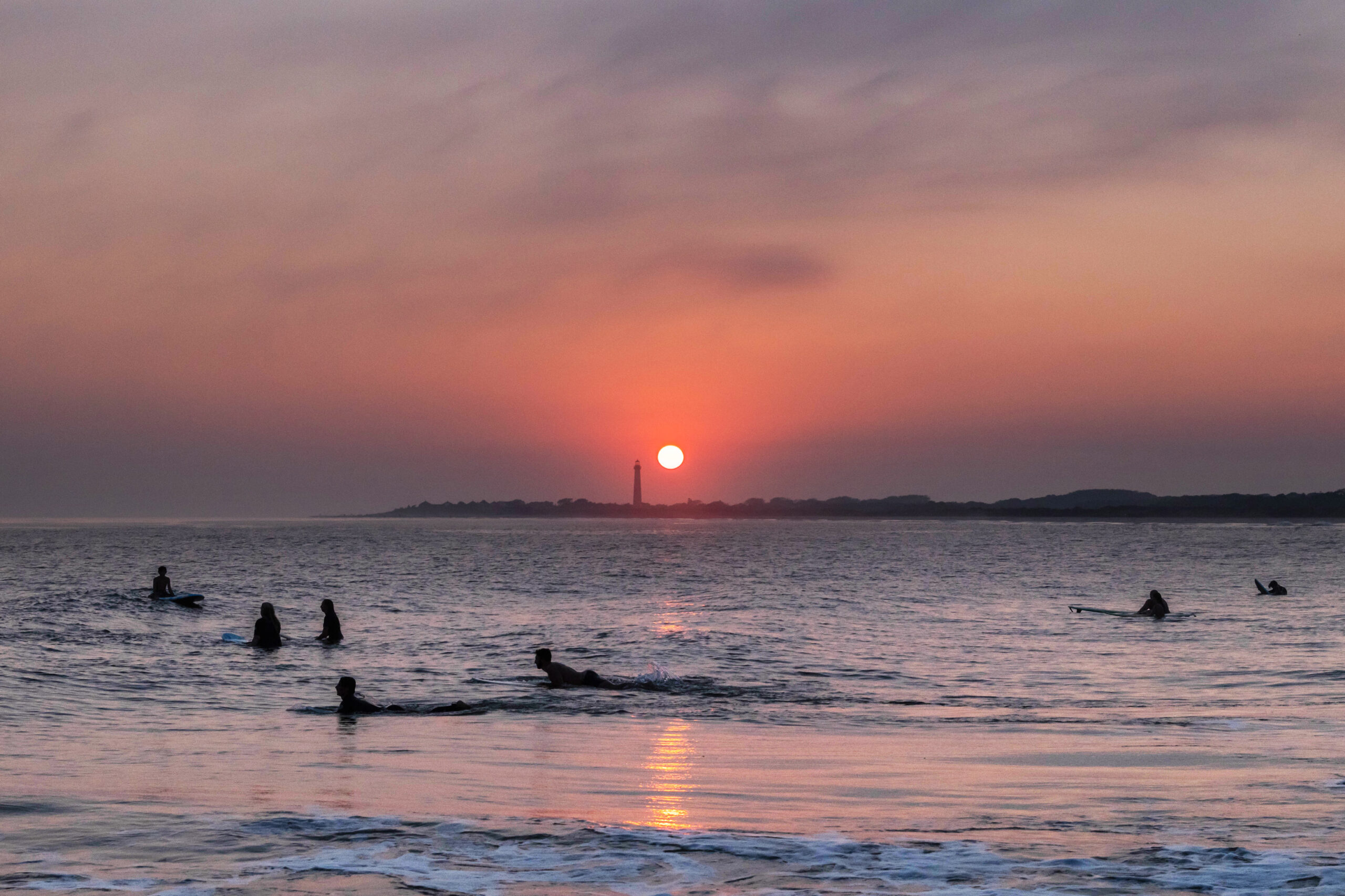 A wide view of sunset at the ocean with the Cape May Lighthouse at the horizon line. There are a group of surfers silhouetted in the ocean. The sun is bright orange pink, and the sky is pink, orange, and purple with purple haze at the horizon.