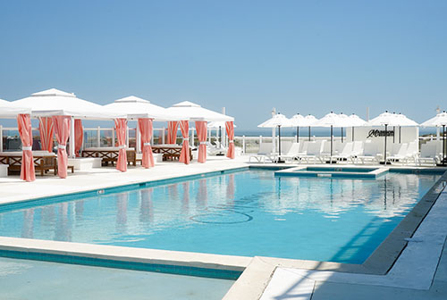 Swimming pool surrounded by white and peach cabanas, and white umbrellas and lounge chairs