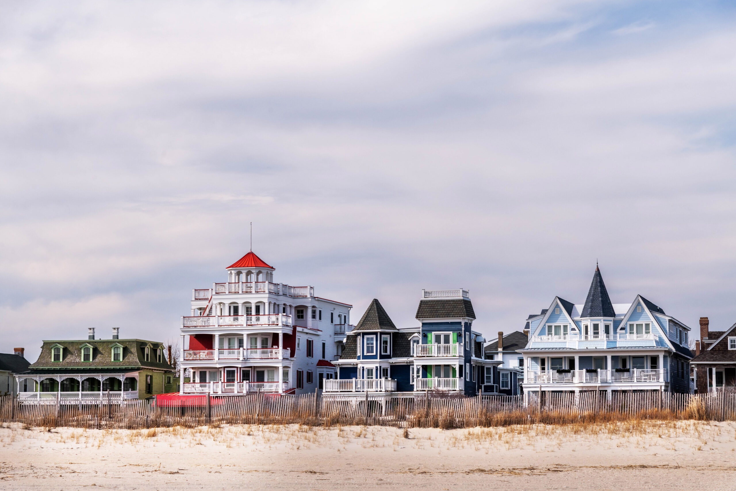 A wide view of four Victorian houses behind the beach. There are wispy white clouds in the sky, and the sun is shining on the houses.