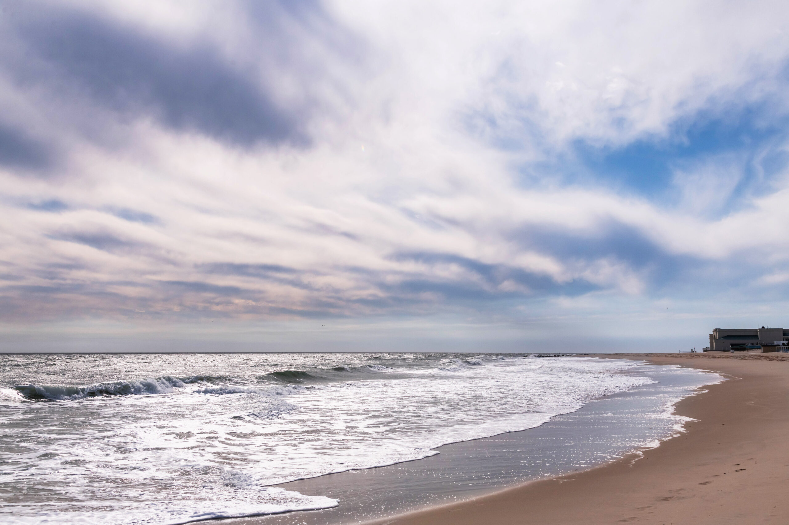 A wide view of the beach, sky, and ocean. There are thin white and gray clouds in the blue sky. The ocean looks silver, and Convention Hall can be seen in the distance.