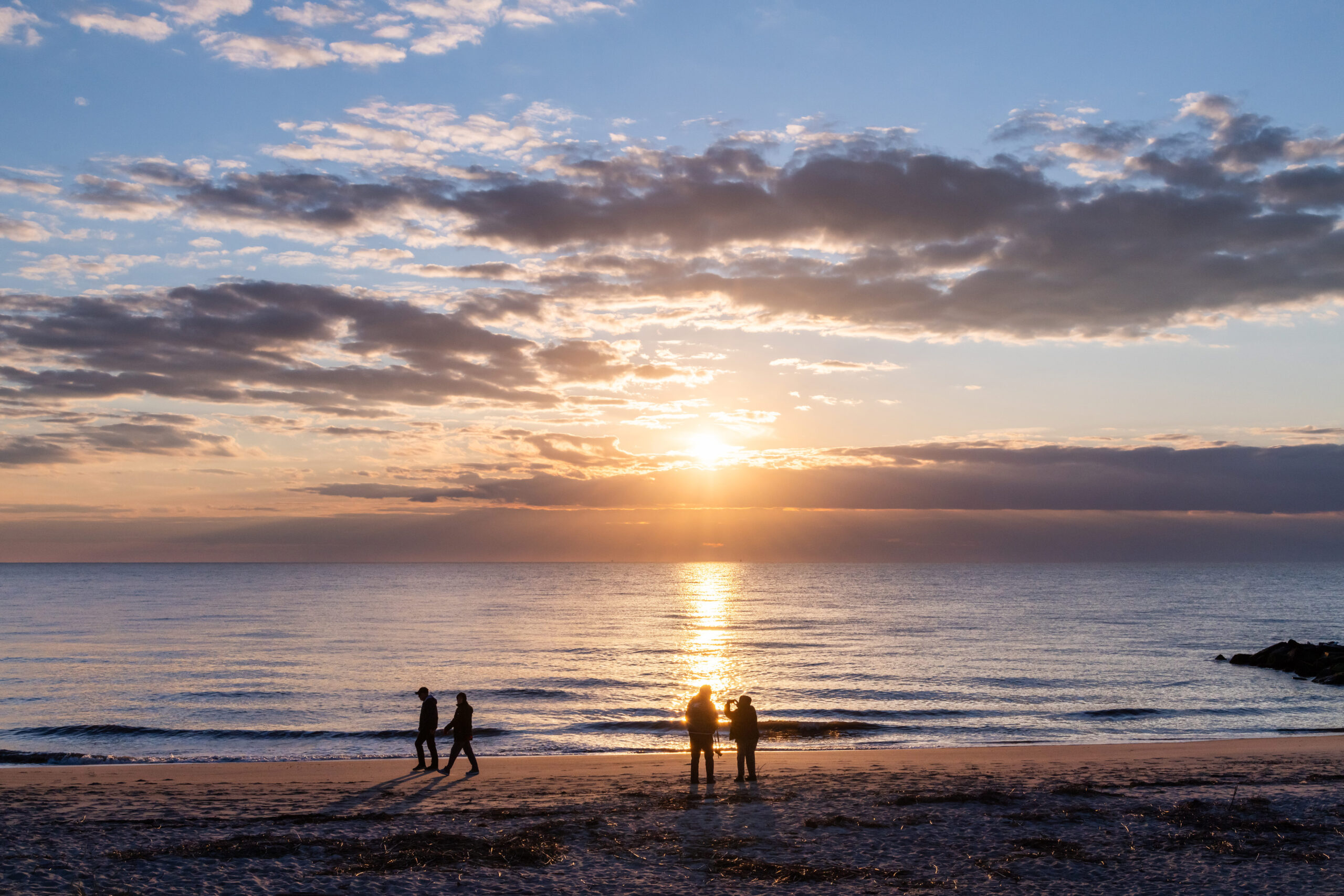 One couple walking on the beach and another couple watching the sun set. There are a few thin clouds in the sky.
