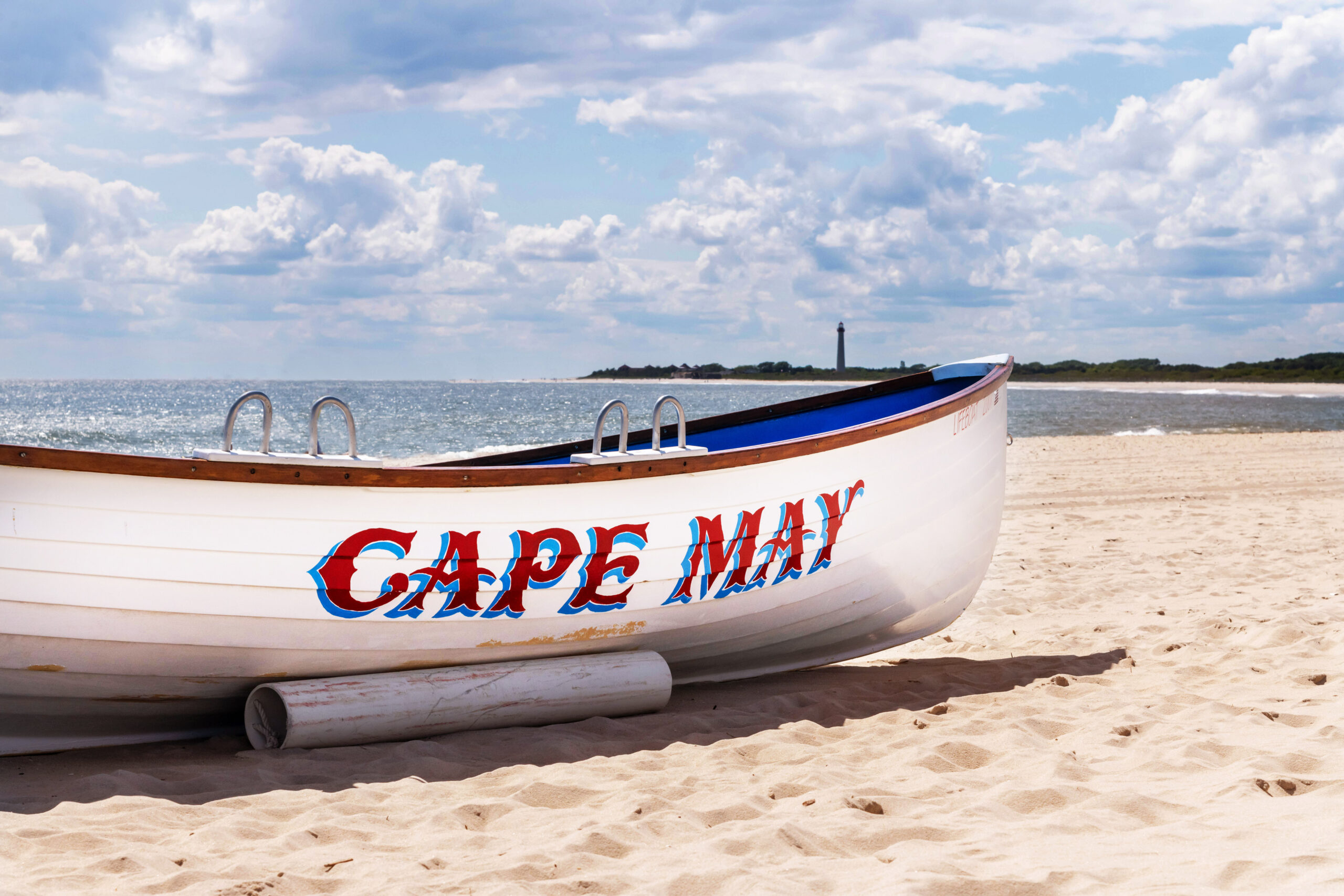 A close up view of a Cape May lifeguard boat on the beach with the Cape May Lighthouse in the distance. The boat is white with "CAPE MAY" written in red letters with blue outline. The sky is blue with puffy white and blue clouds.