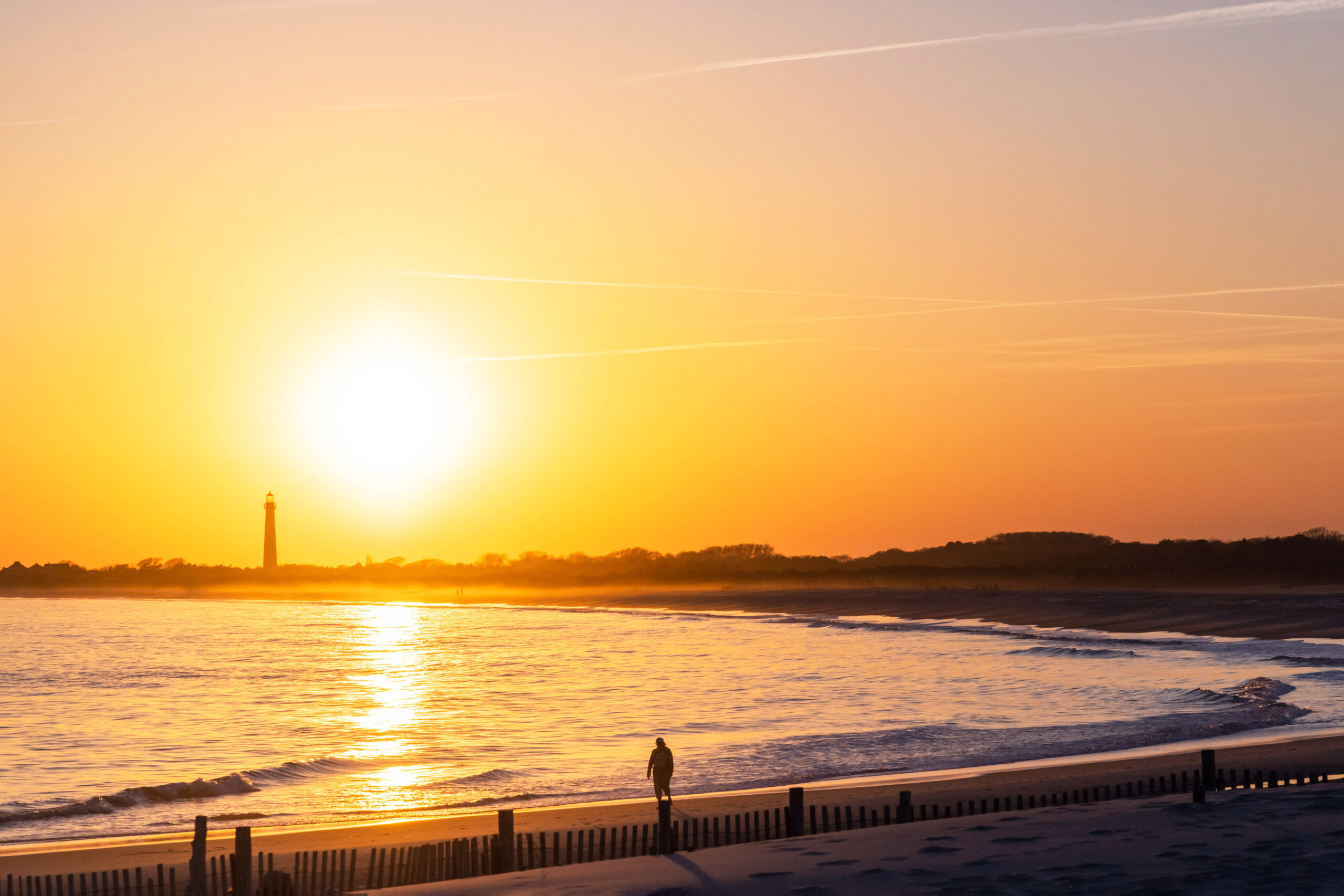 A wide view of a person walking along the ocean at sunset with the Cape May Lighthouse in the background. The sky is bright orange and yellow, and the colors are reflected in the calm ocean.