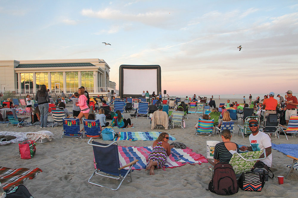 An inflatable screen set up next to Convention Hall in Cape May. A crowd of people on beach chairs and towels are spread out in front of it