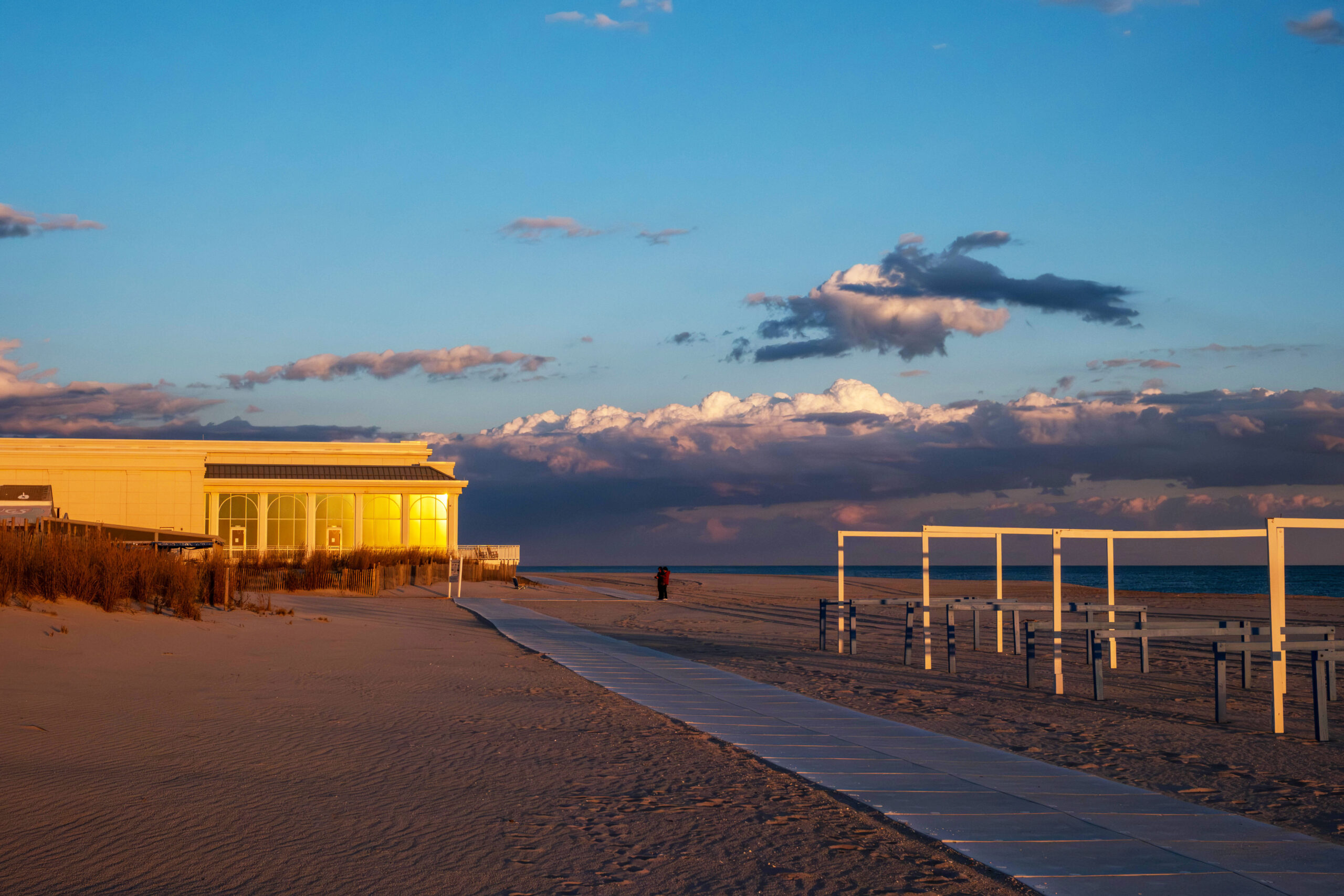 A wide view of the Convention Hall and the beach at sunset. Sunlight is shining on Convention Hall and bare beach cabanas. There are a few dark purple, white, and pink puffy clouds at the horizon.