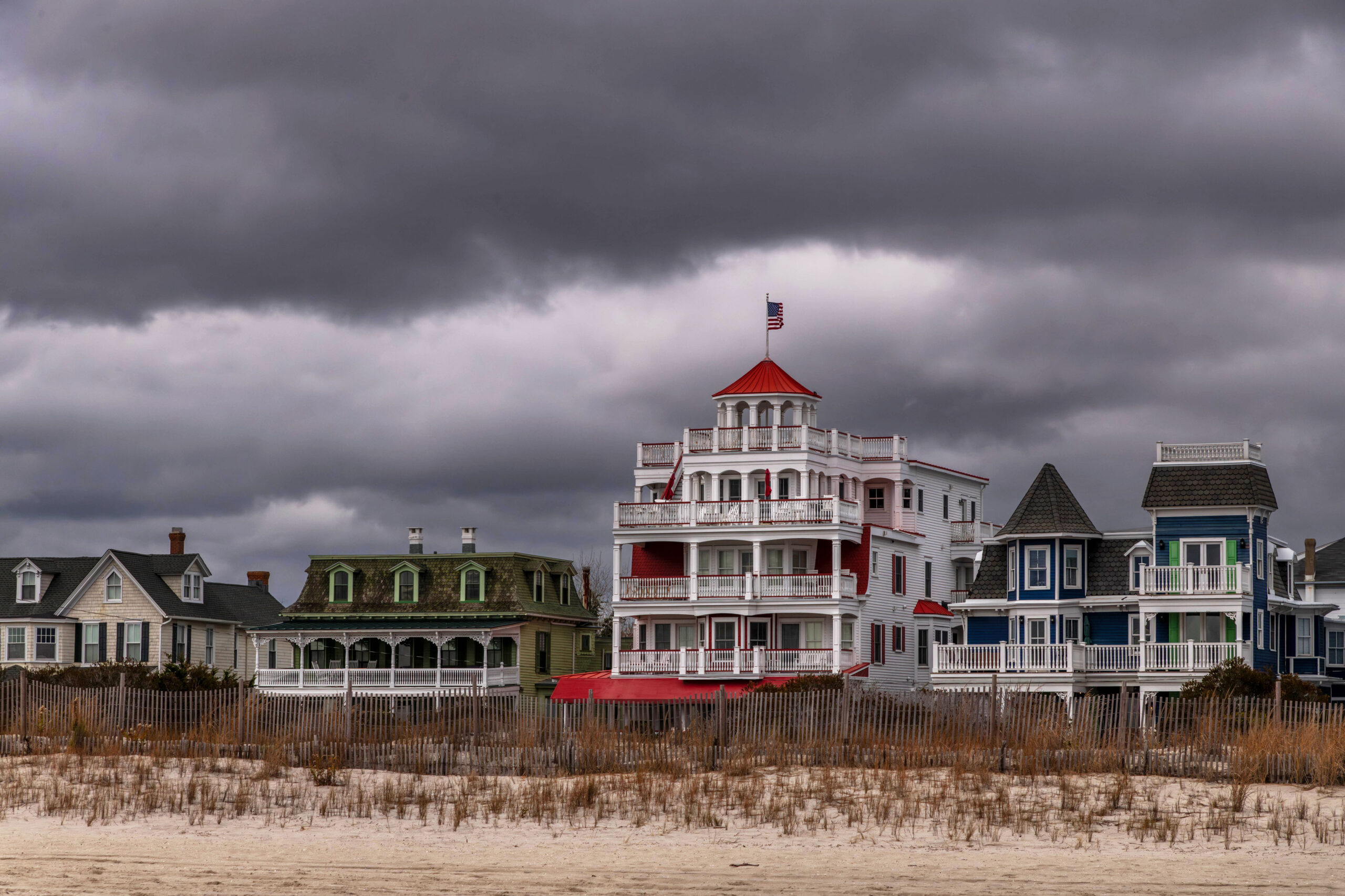 A wide view of four Victorian homes behind the beach with dark gray and white puffy clouds in the sky. The home on the far left is tan. The home to the right of it is green. The home in the middle is white with red shudders. The home on the far right is blue with green shudders.