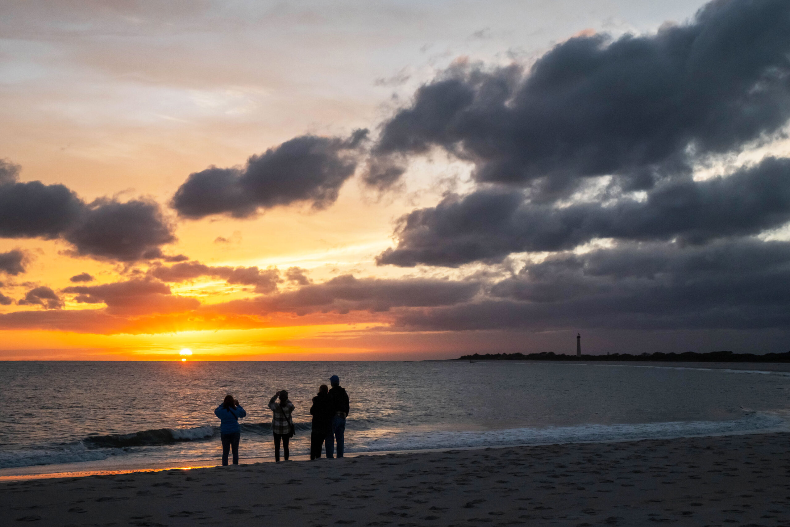 A wide view of a group of people on the beach at sunset. The Cape May Lighthouse is in the distance on the horizon. There are a few puffy dark clouds in the sky. The sky is orange and pink.