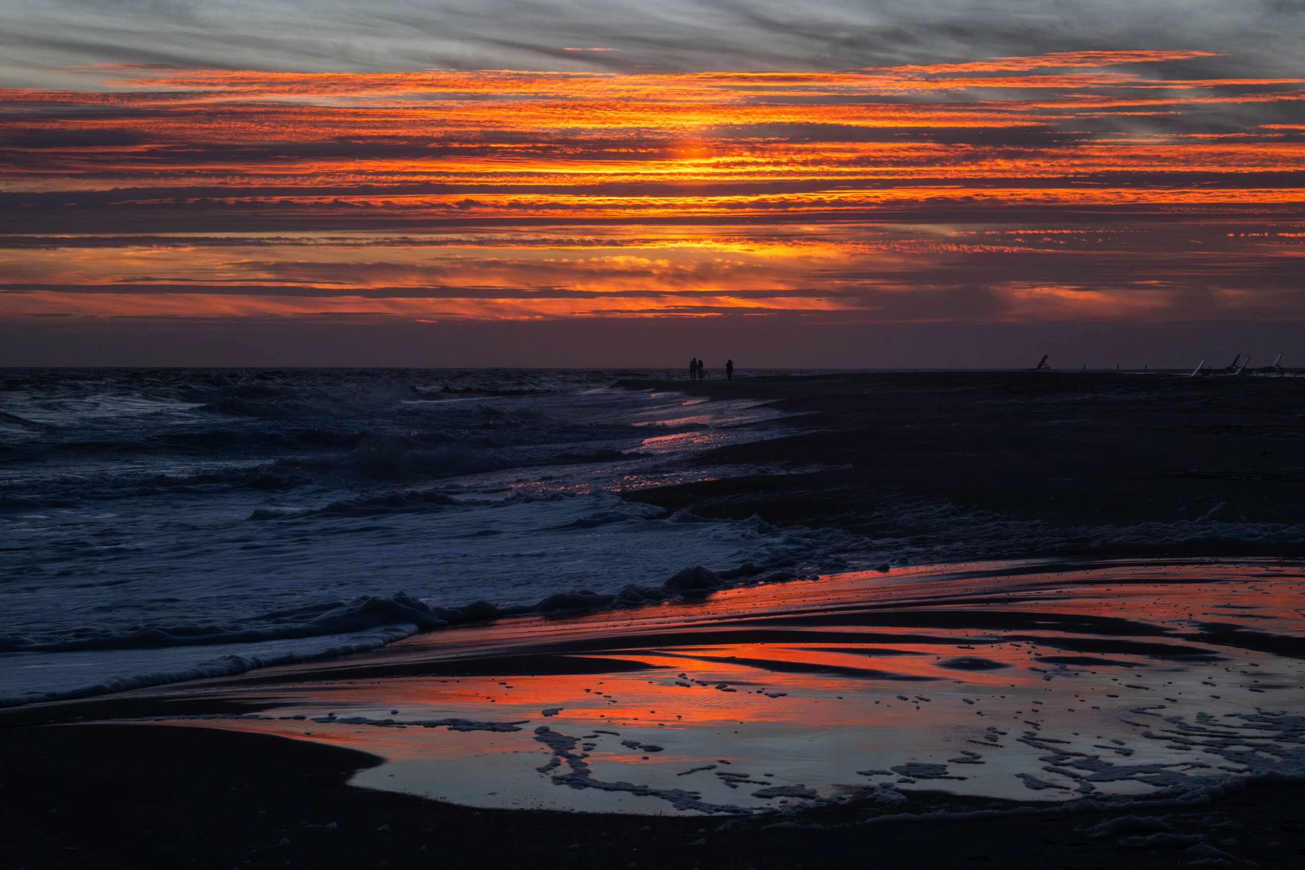 A wide view of a colorful sunset at the beach. The sky has stripes of bright orange and thin purple clouds. The colors from the clouds are reflected in the shoreline on the beach.