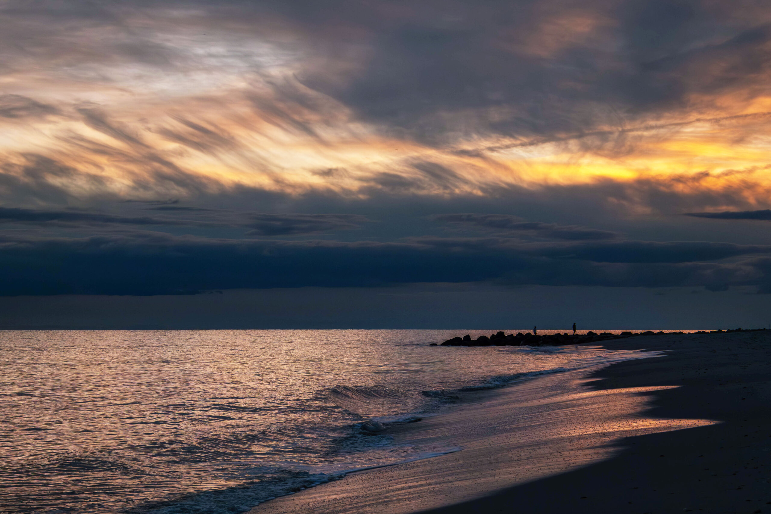 A wide view of the beach and ocean at sunset. There are thin dark purple and yellow clouds in the sky. The colors are reflected in the ocean that looks bright. The sand is dark.