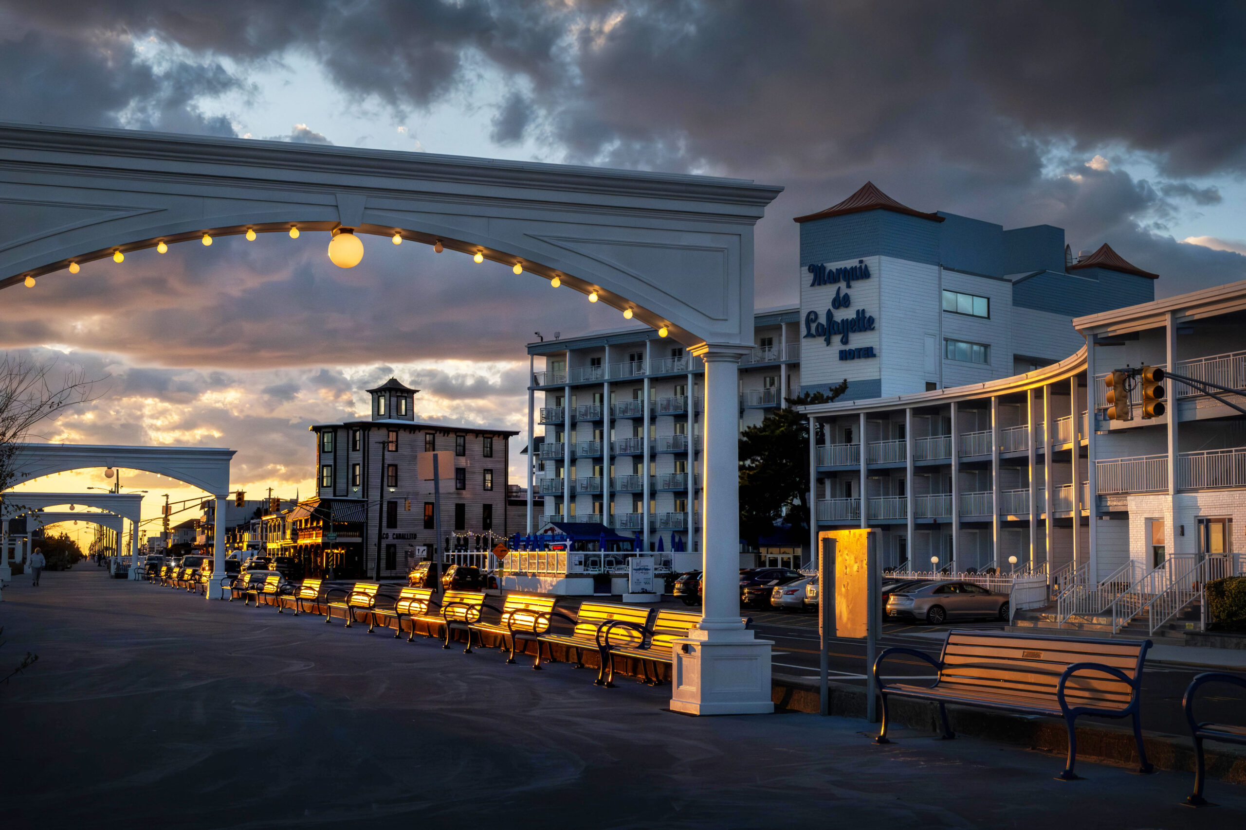 A wide view of the Cape May arches on the promenade with the Marquis de Lafayette hotel in the background at sunset. Sunlight is shining on the benches on the promenade, and there are dark purple and pink puffy clouds in the sky.