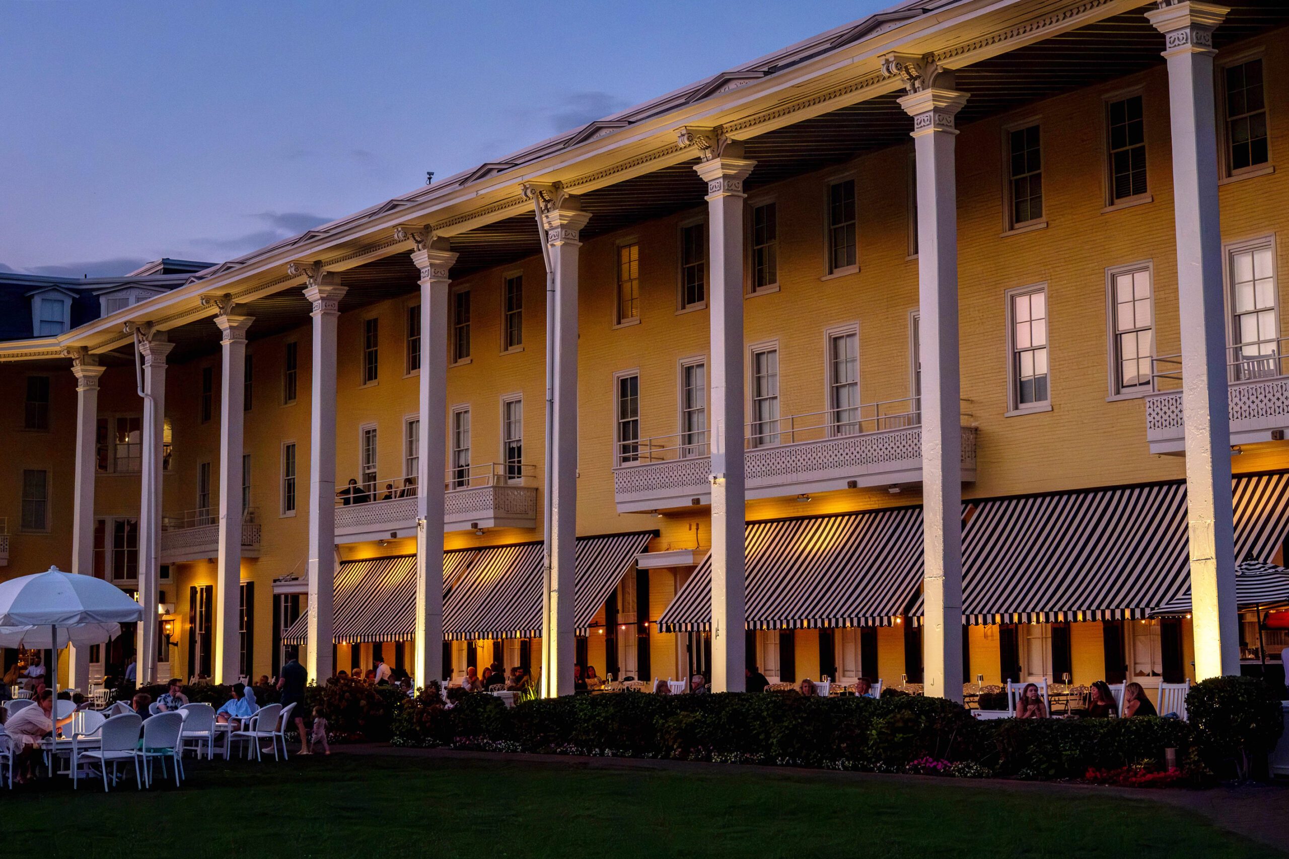 A wide view of Congress Hall in the early evening. Lights are shining under black and white awnings, and there are a few white chairs and a white umbrella in the corner on the front lawn.