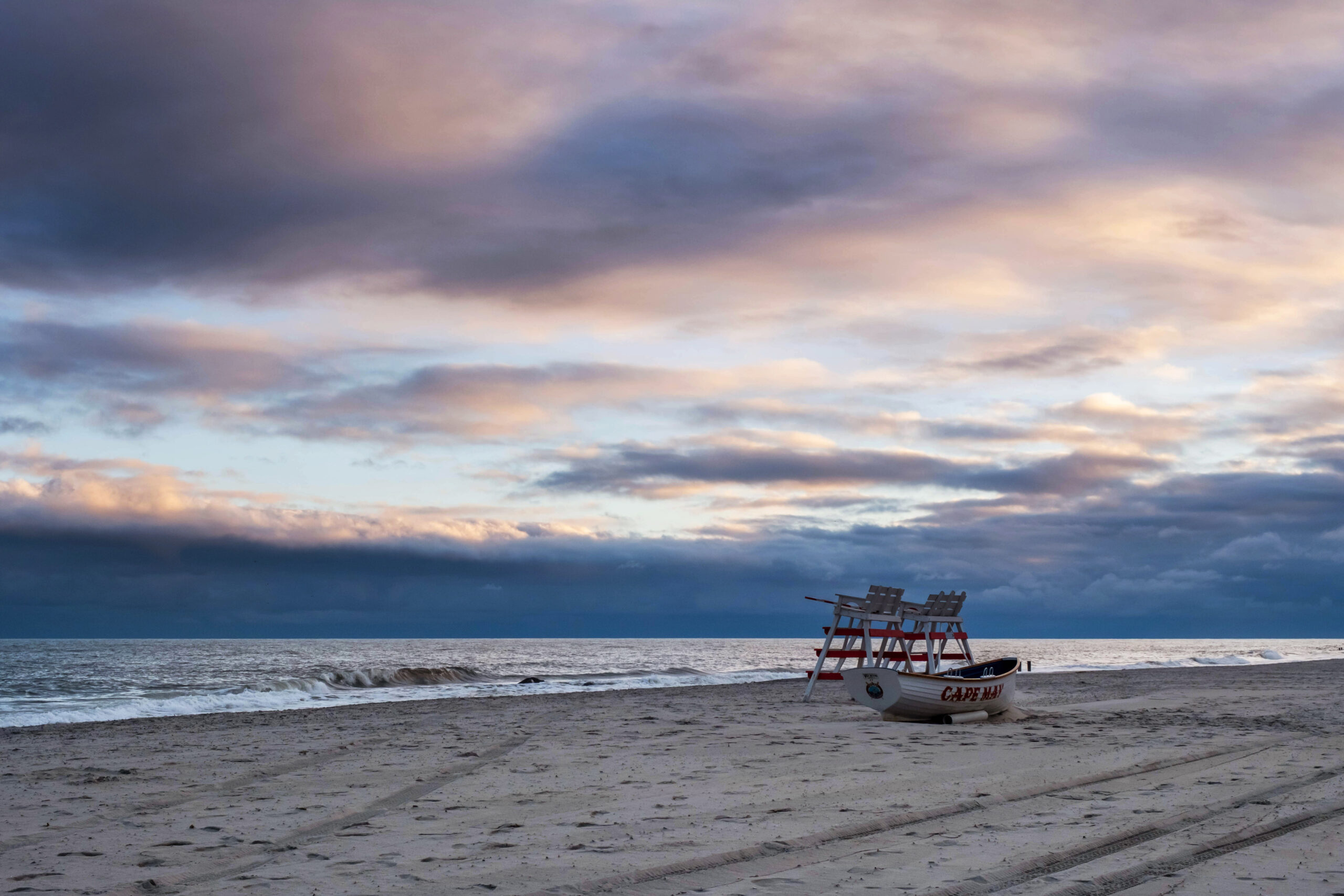 A wide view of lifeguard stands and a Cape May lifeguard boat on the beach with colorful orange and purple clouds.