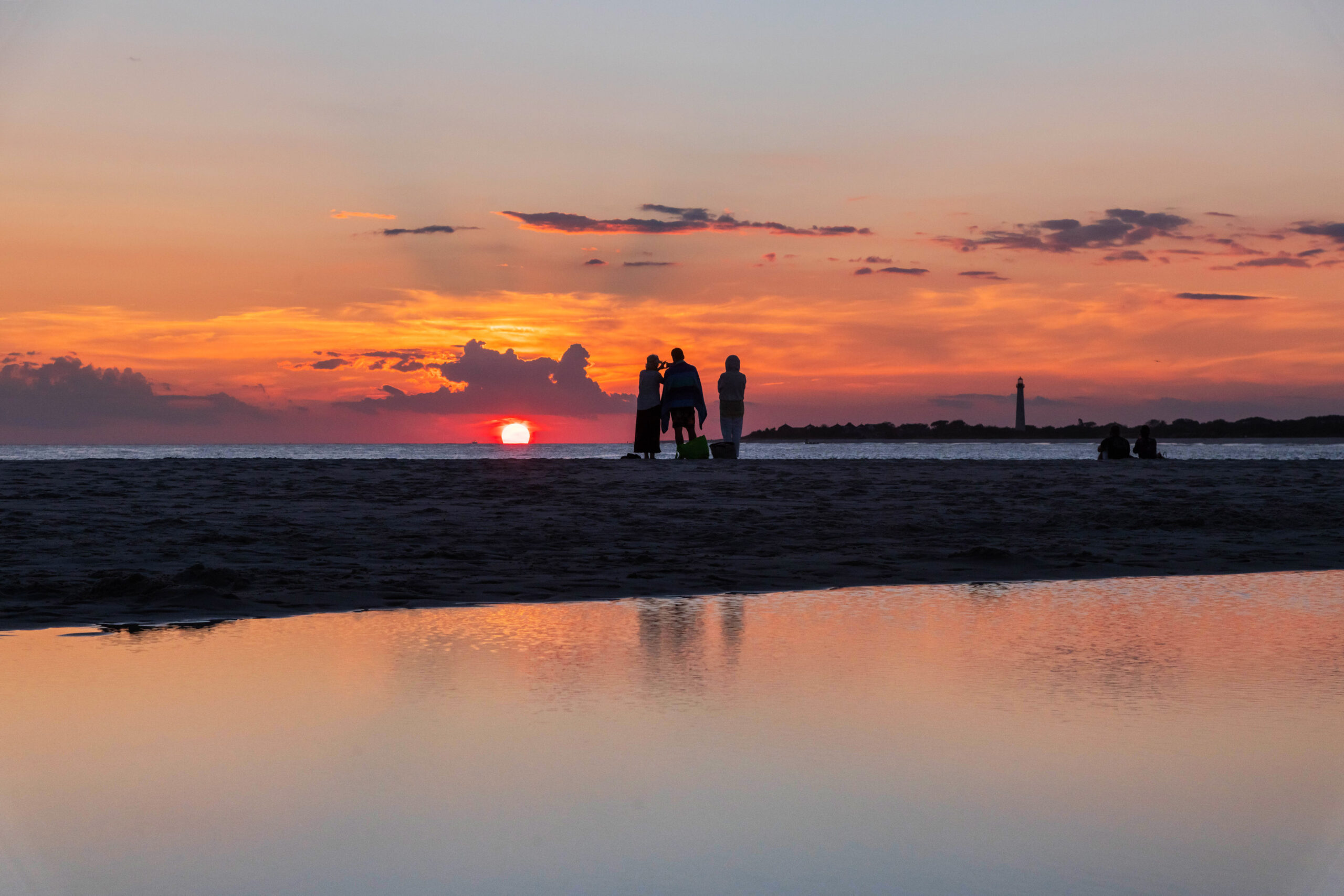 Three people standing at the edge of the beach watching the sunset with the Cape May lighthouse in the distance. The sun is bright pink, and there are purple and orange clouds in the sky. There is a body of water in the foreground, and the colors in the sky are reflected in the water.