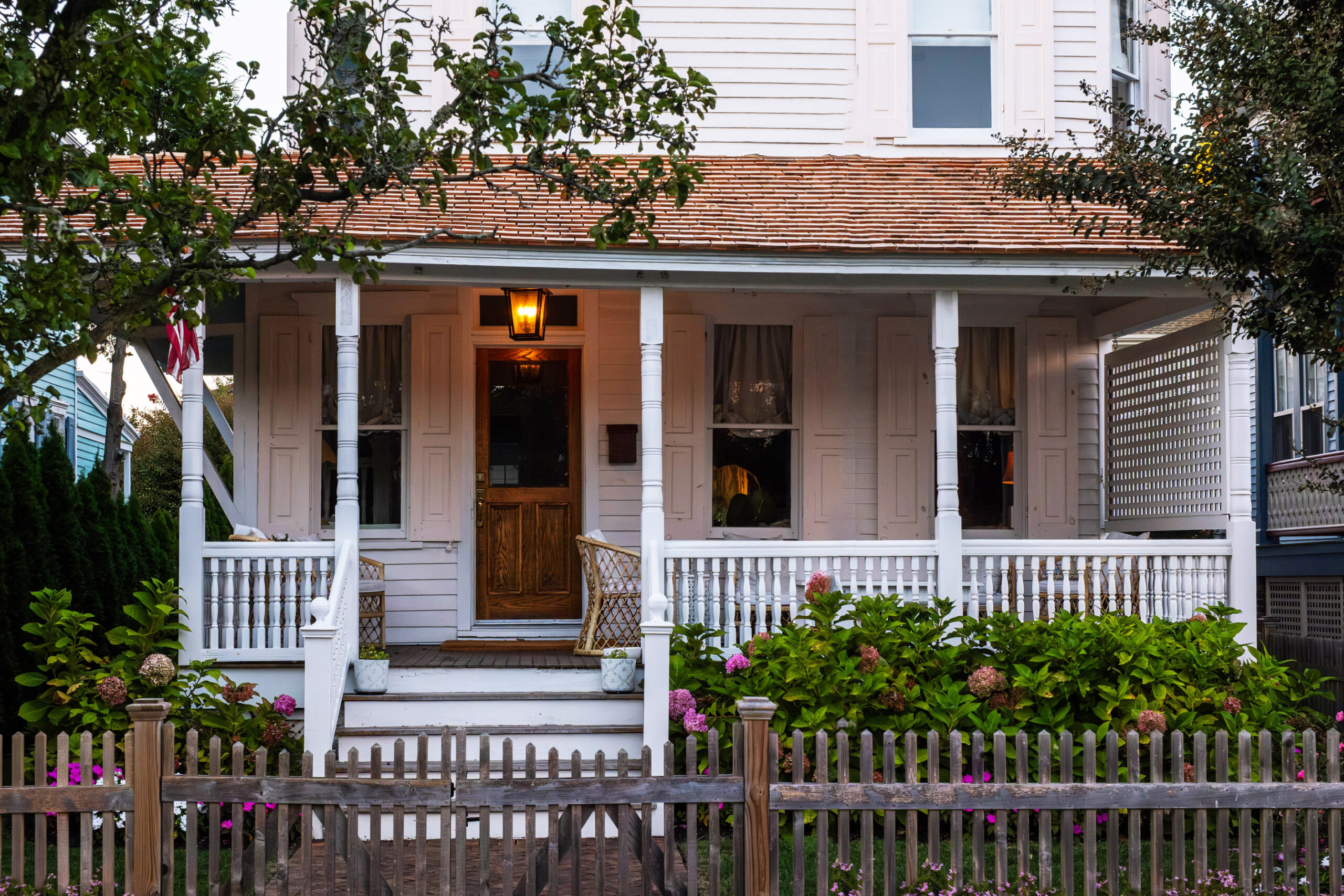 A front porch of a Victorian home. The porch is white with a brown down. There is a light on in the front porch in from of the door. There are a few pink hydrangea in some bushes in front of the porch. There is a brown picket fence in front of the house.